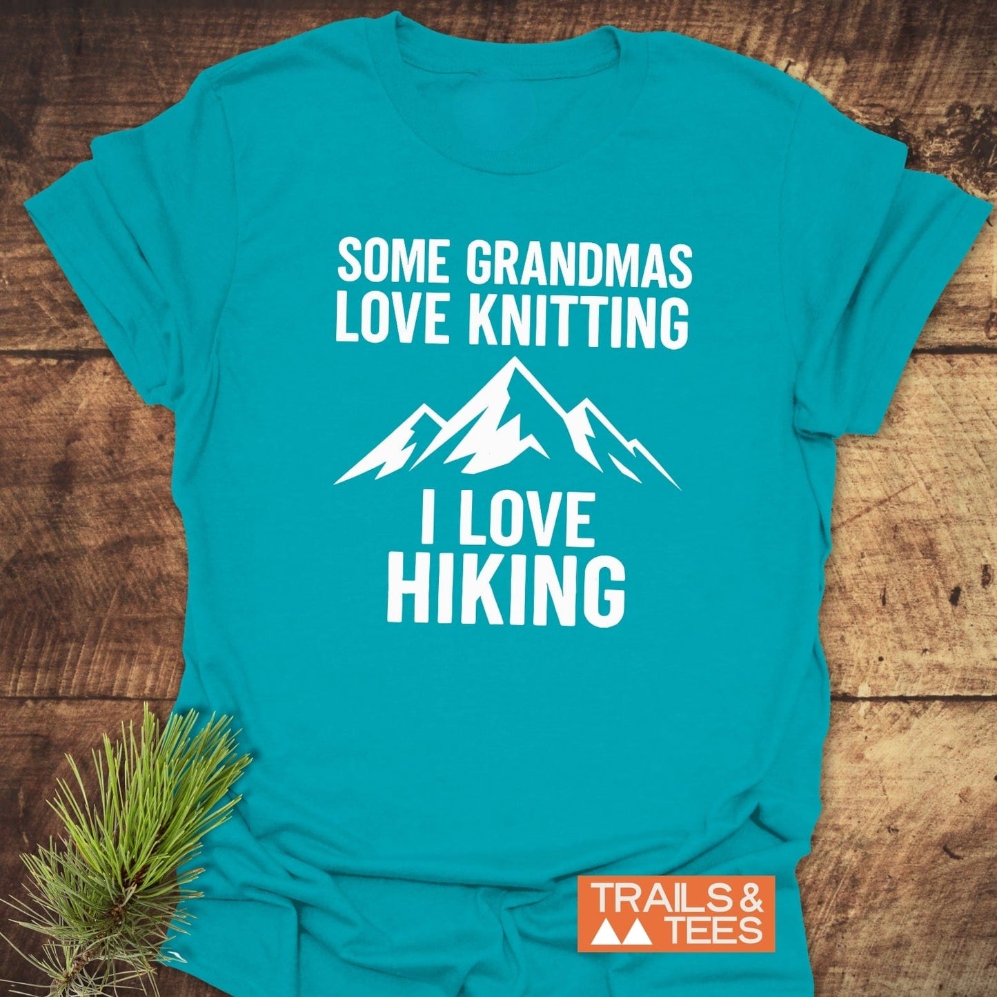 Hiking Grandma Funny T-Shirt with a white mountain graphic and slogan, laid flat with a pine sprig, showcasing its rustic, outdoor-inspired design.