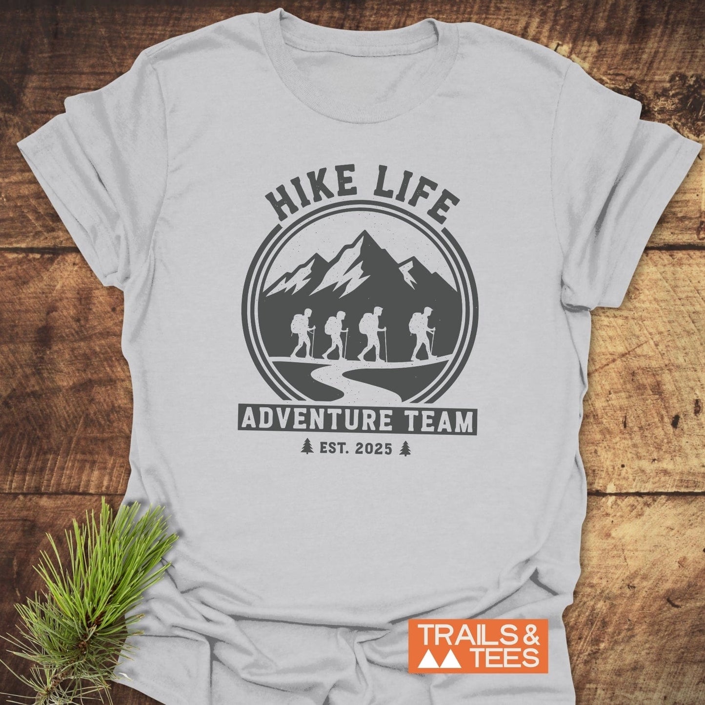 Adventure Team T-Shirt flat on wood, features a circular graphic with backpackers, HIKE LIFE text, and TRAILS & TEES logo, emphasizing outdoor adventure spirit.