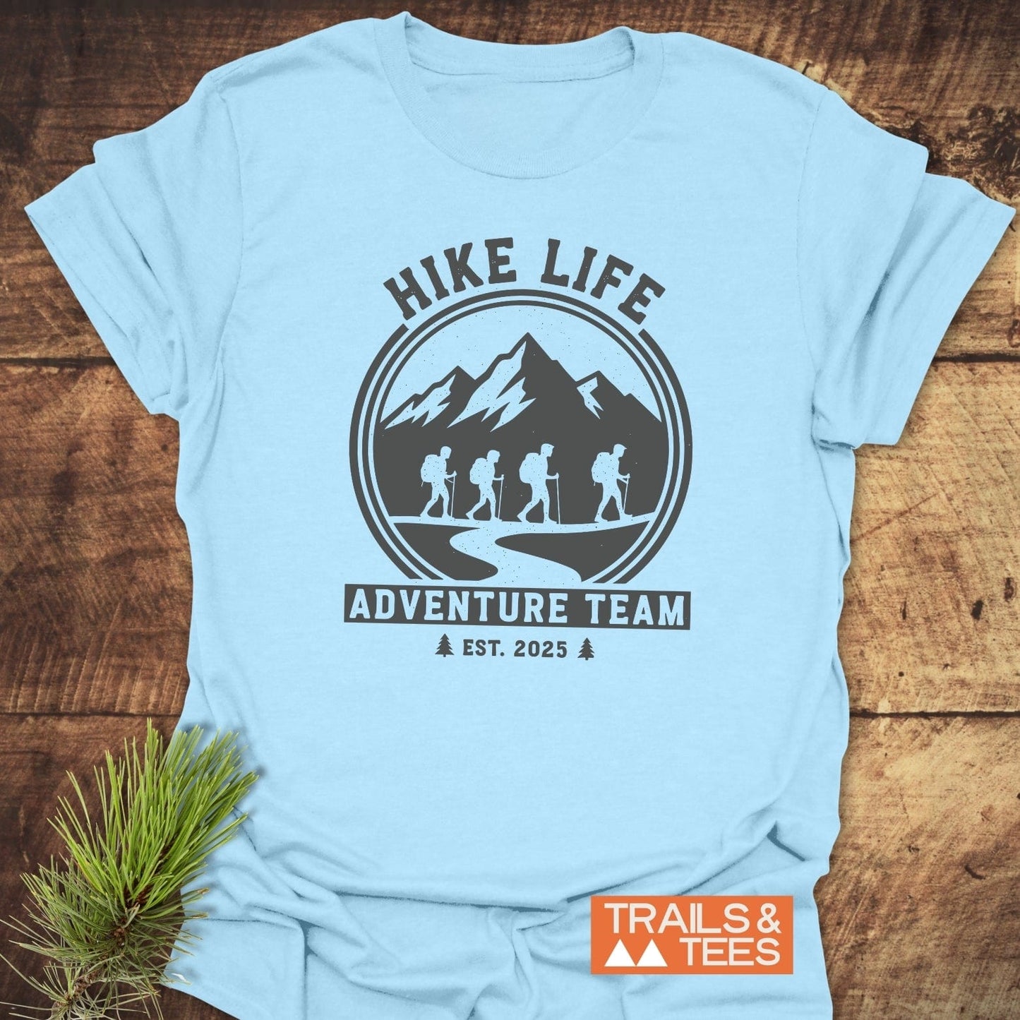 Adventure Team T-Shirt in light blue, featuring a badge-style graphic with hikers and mountains, emphasizing comfort, durability, and eco-friendly production.