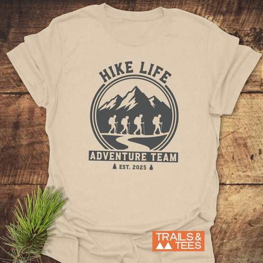 Adventure Team T-Shirt featuring a mountain graphic with hikers, HIKE LIFE, and ADVENTURE TEAM text, laid flat on a wooden surface, next to pine sprig.