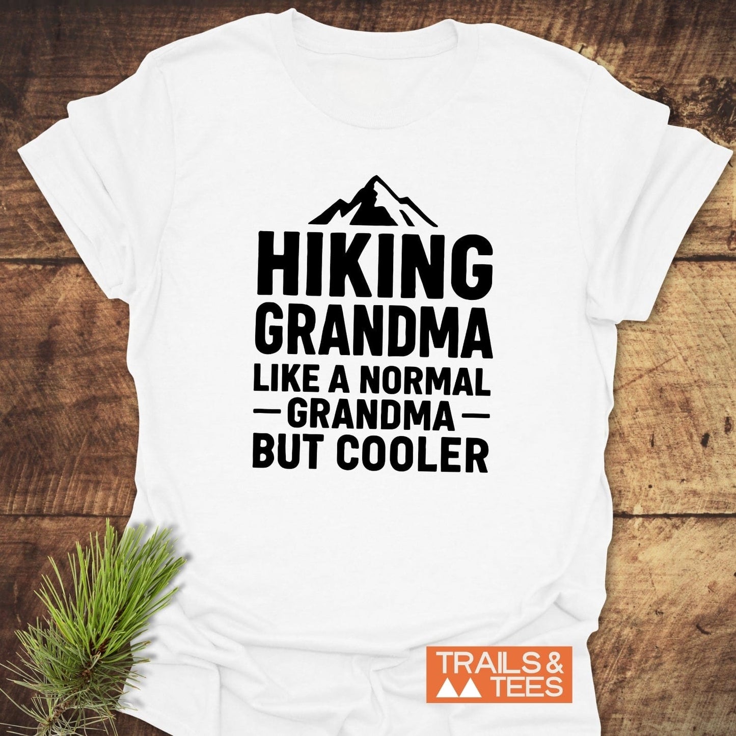 Hiking Grandma T-Shirt featuring a mountain silhouette with bold text, displayed on wood with a pine sprig and Trails & Tees logo.