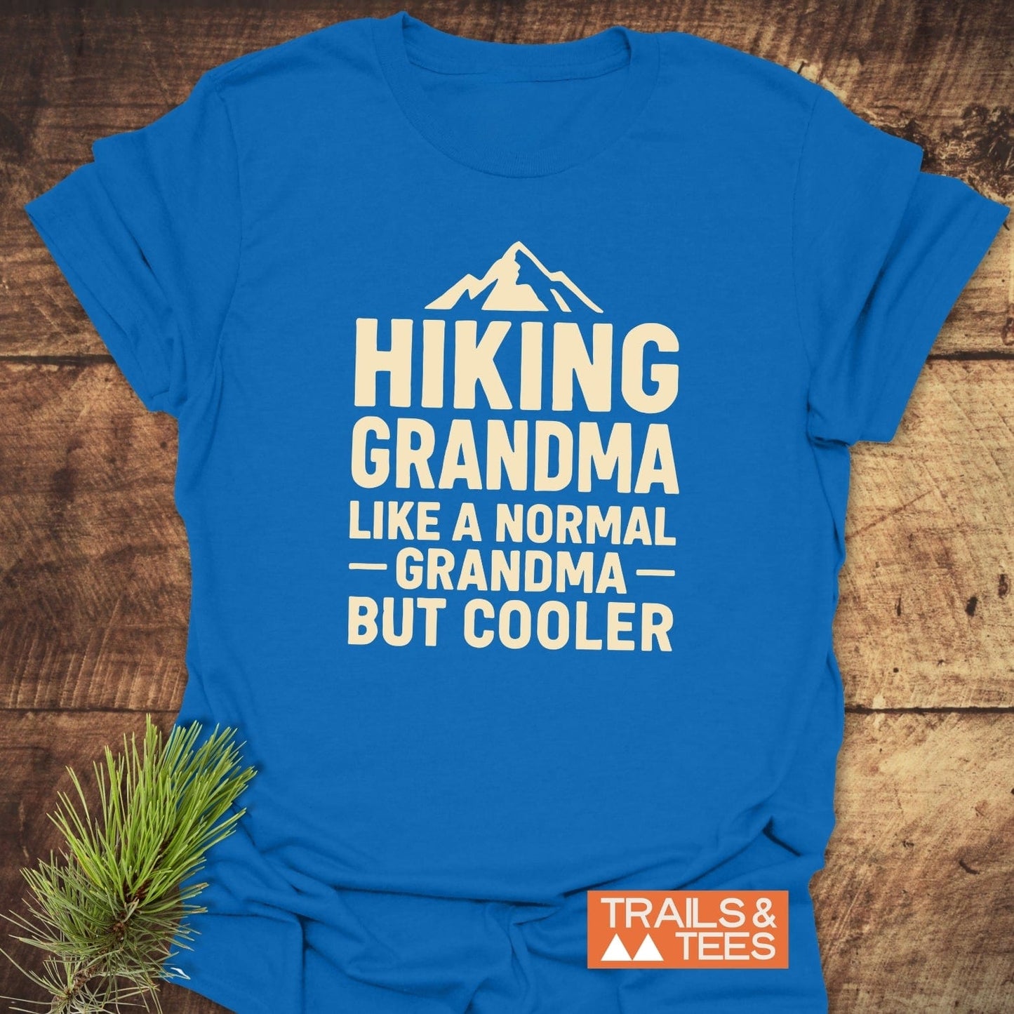 Hiking Grandma T-Shirt with mountain graphic, laid on wood. Cream print: “Hiking Grandma, Like a Normal Grandma, But Cooler.” Includes Trails & Tees logo.