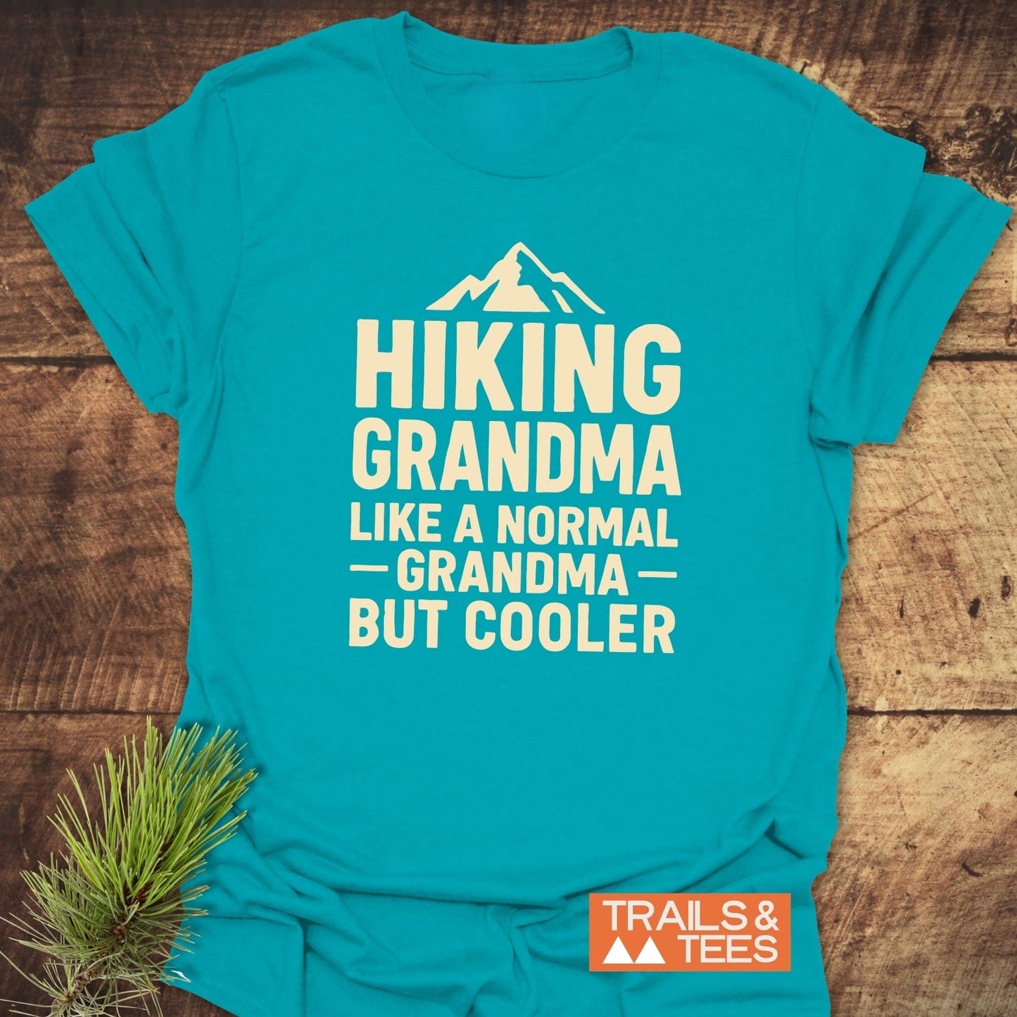 Turquoise T-shirt with “HIKING GRANDMA” print, featuring a mountain silhouette. Includes Trails & Tees logo and pine sprig accent for rustic appeal.