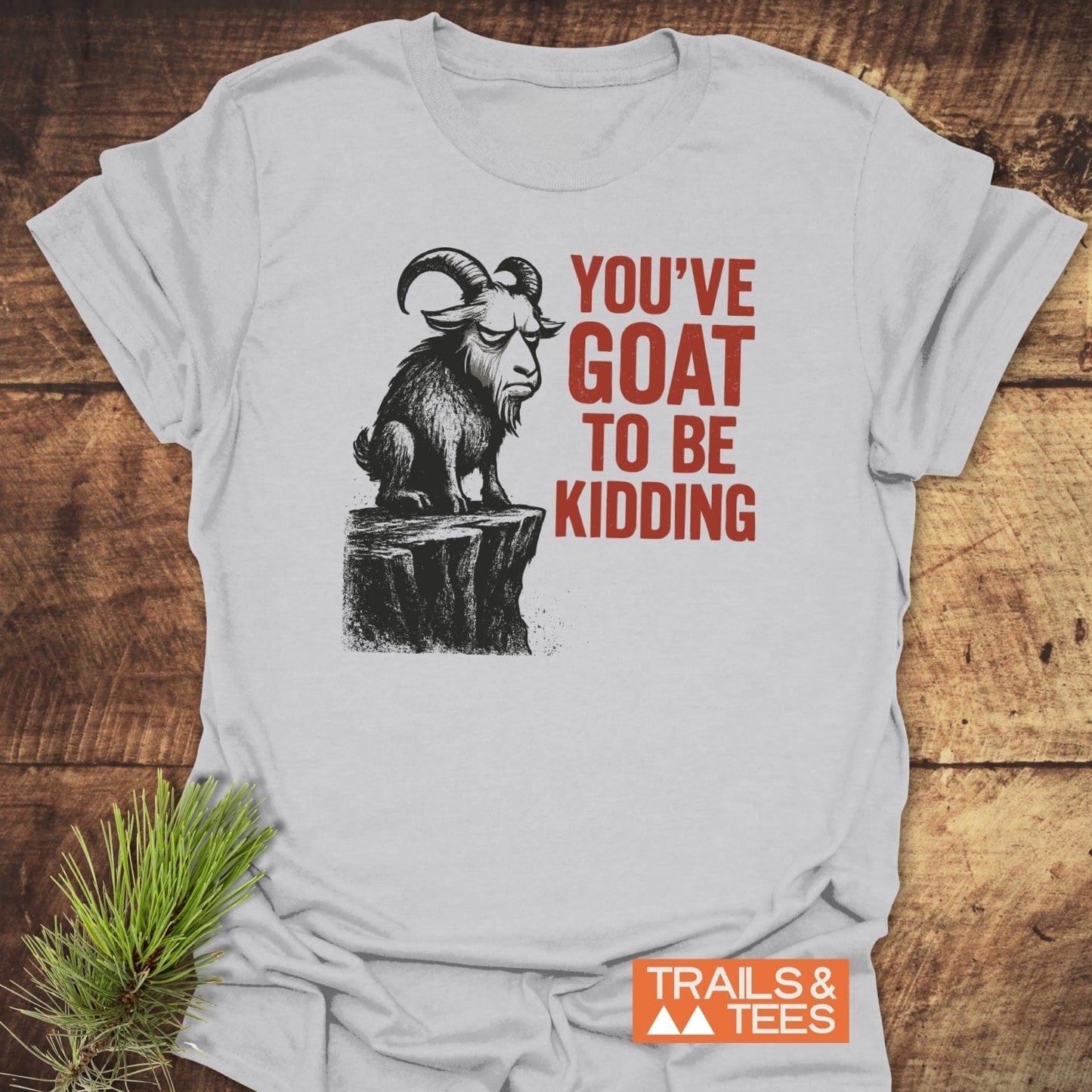 Kidding Goat Funny T-Shirt with a grumpy goat illustration and bold text, laid on wood, highlighting its soft cotton and durable print.