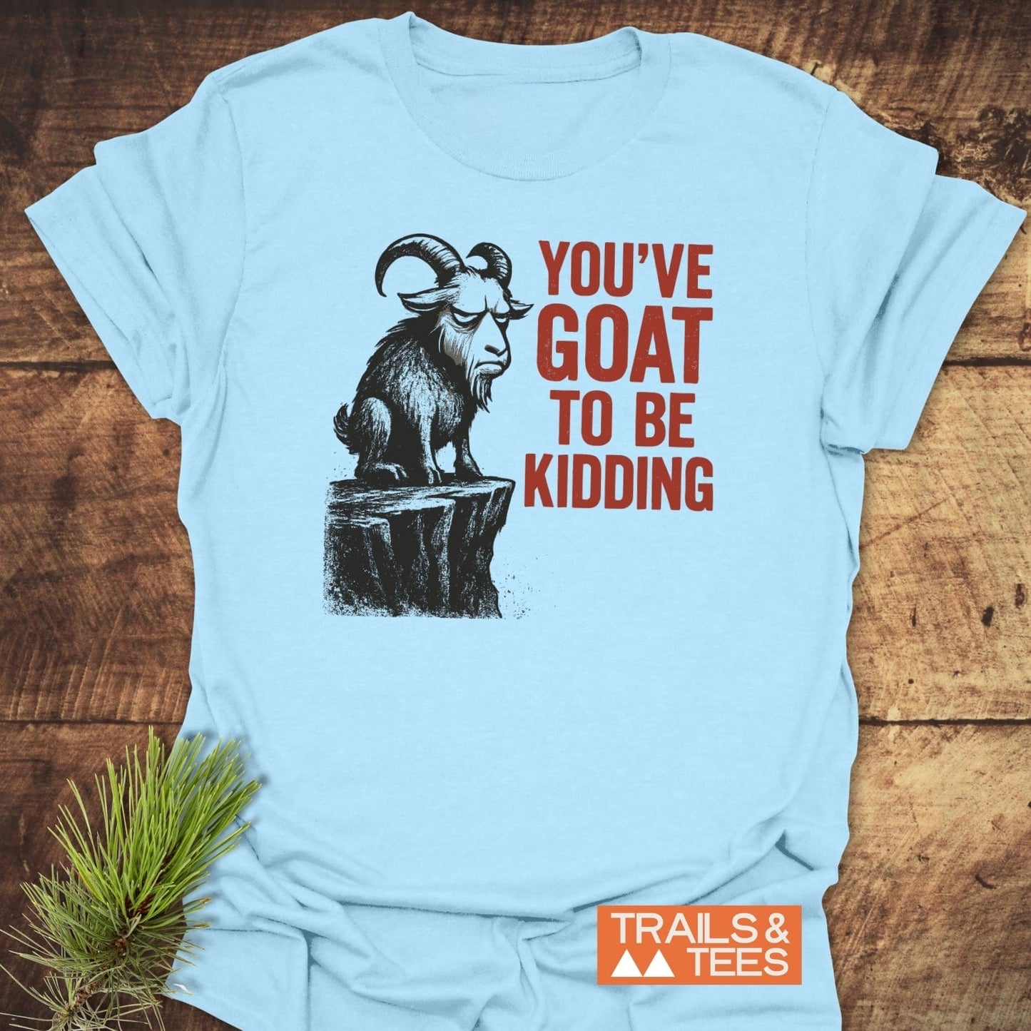 Kidding Goat Funny T-Shirt features a grumpy goat on a rocky ledge with “YOU’VE GOAT TO BE KIDDING” slogan, emphasizing comfort and durability from Trails & Tees.