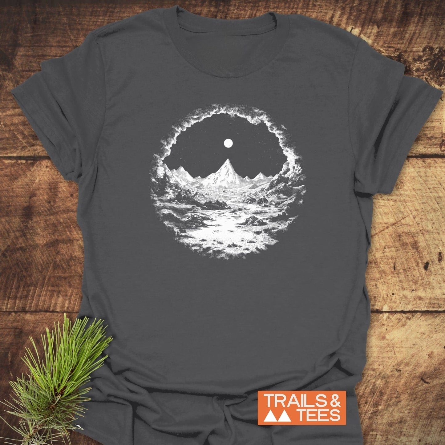 Nighttime Wandering T-Shirt in charcoal-gray with a white mountain graphic, showcasing durable design and comfort, laid on wood with a pine sprig.
