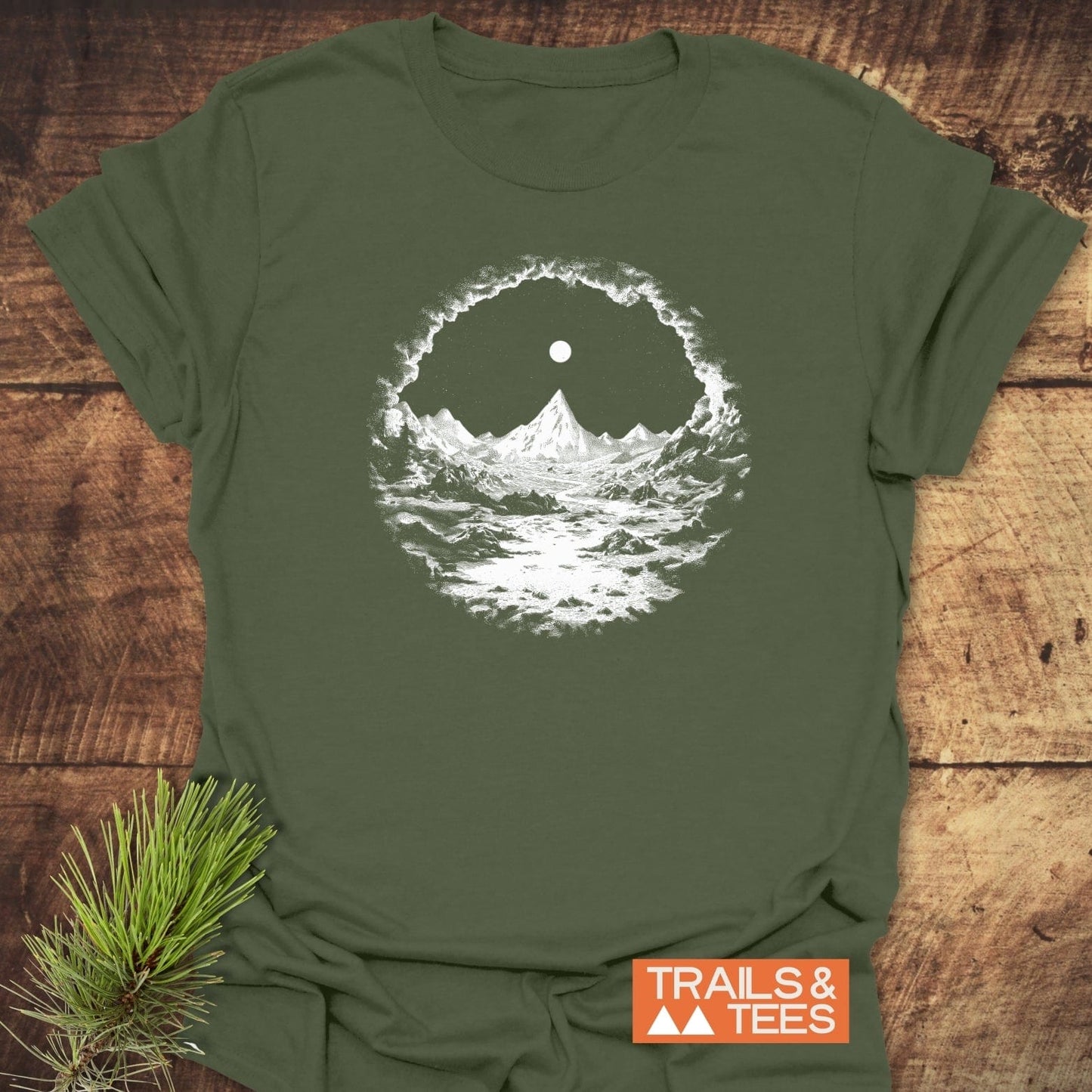 Nighttime Wandering T-Shirt: olive-green tee with white mountain and moon graphic, orange “TRAILS & TEES” logo, and pine sprig on wooden surface.