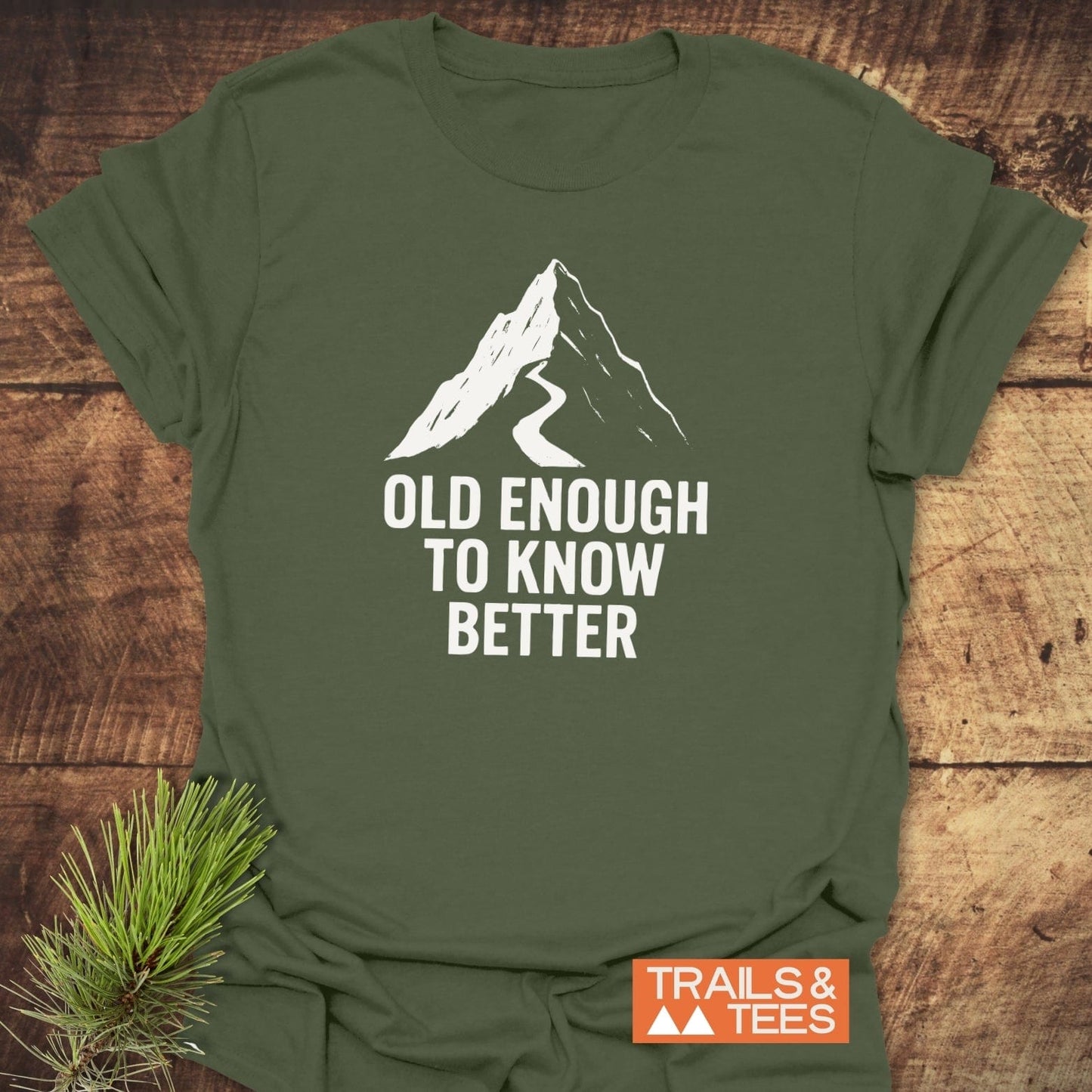 Olive-green T-shirt with mountain silhouette and OLD ENOUGH TO KNOW BETTER slogan. Includes TRAILS & TEES label; styled with pine sprig.