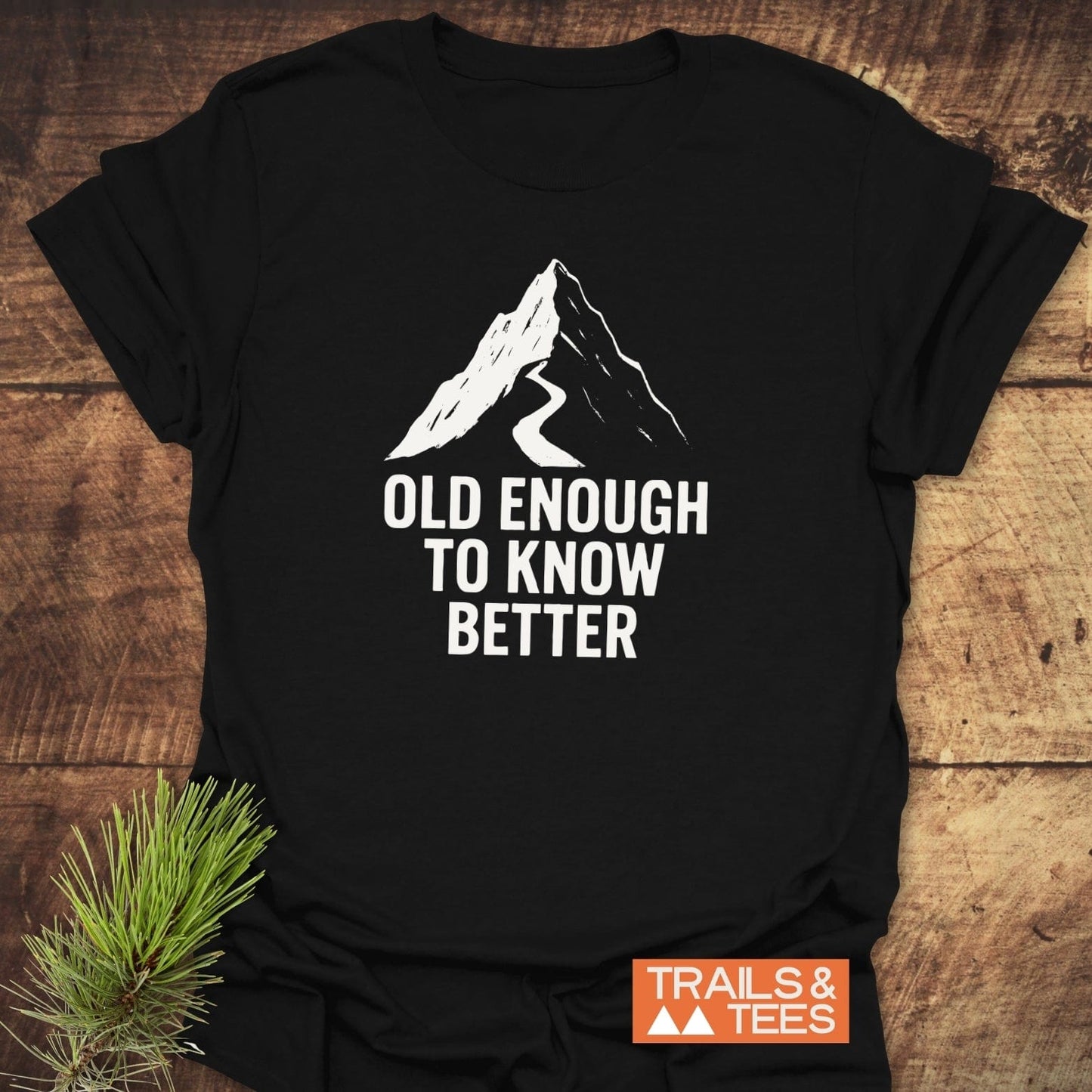Old Enough Funny T-Shirt featuring a stylized mountain peak and slogan, laid on wood. Includes “TRAILS & TEES” logo, embodying outdoor adventure spirit.