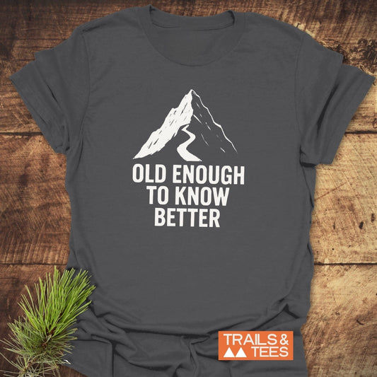 Old Enough Funny T-Shirt with a mountain design and slogan, laid on wood. Crafted from soft cotton for comfort, featuring durable prints and eco-conscious production.