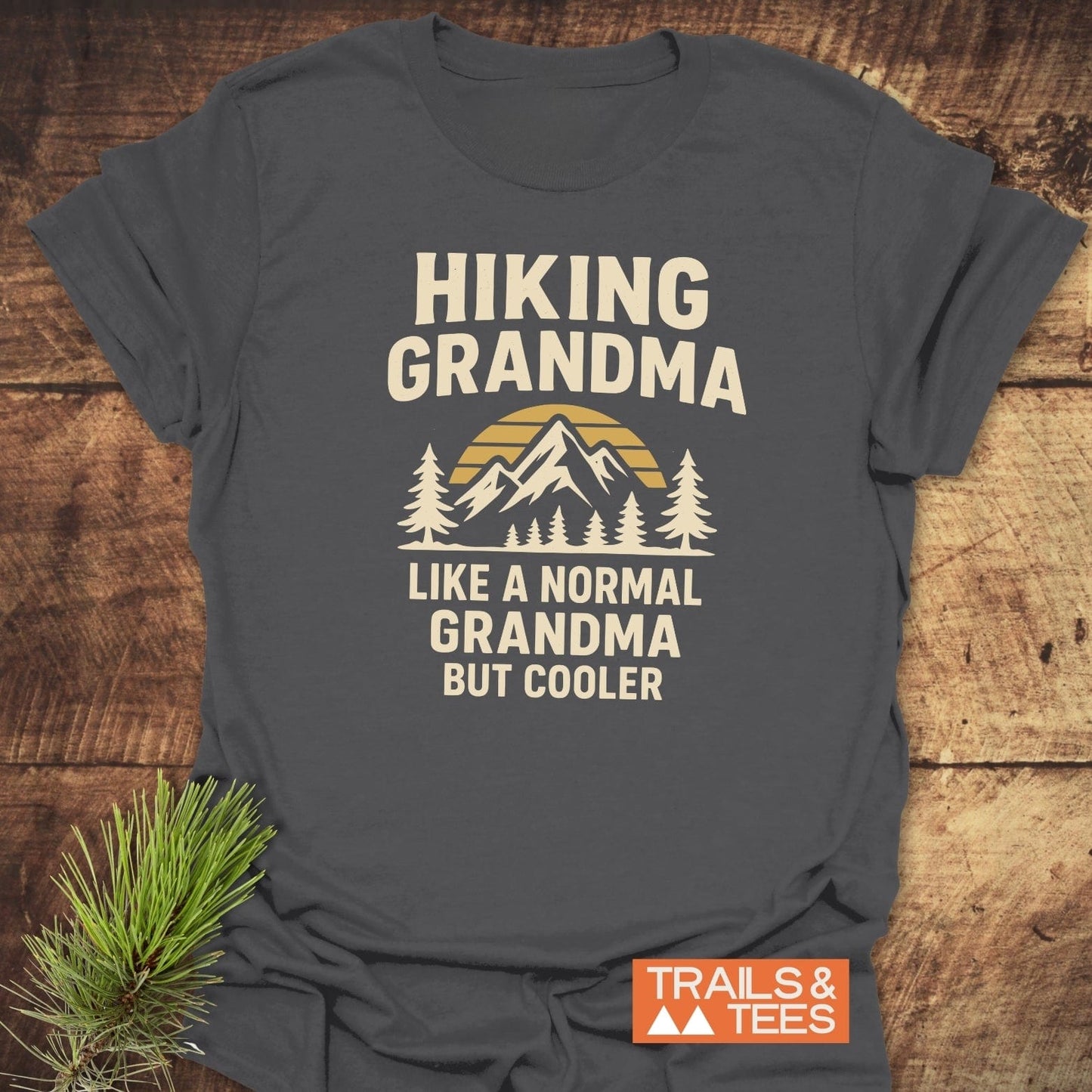 Hiking Grandma Sunrise T-Shirt: dark-gray tee with “HIKING GRANDMA” and mountain graphic. Cream print. Durable, soft cotton, ideal for outdoor enthusiasts.