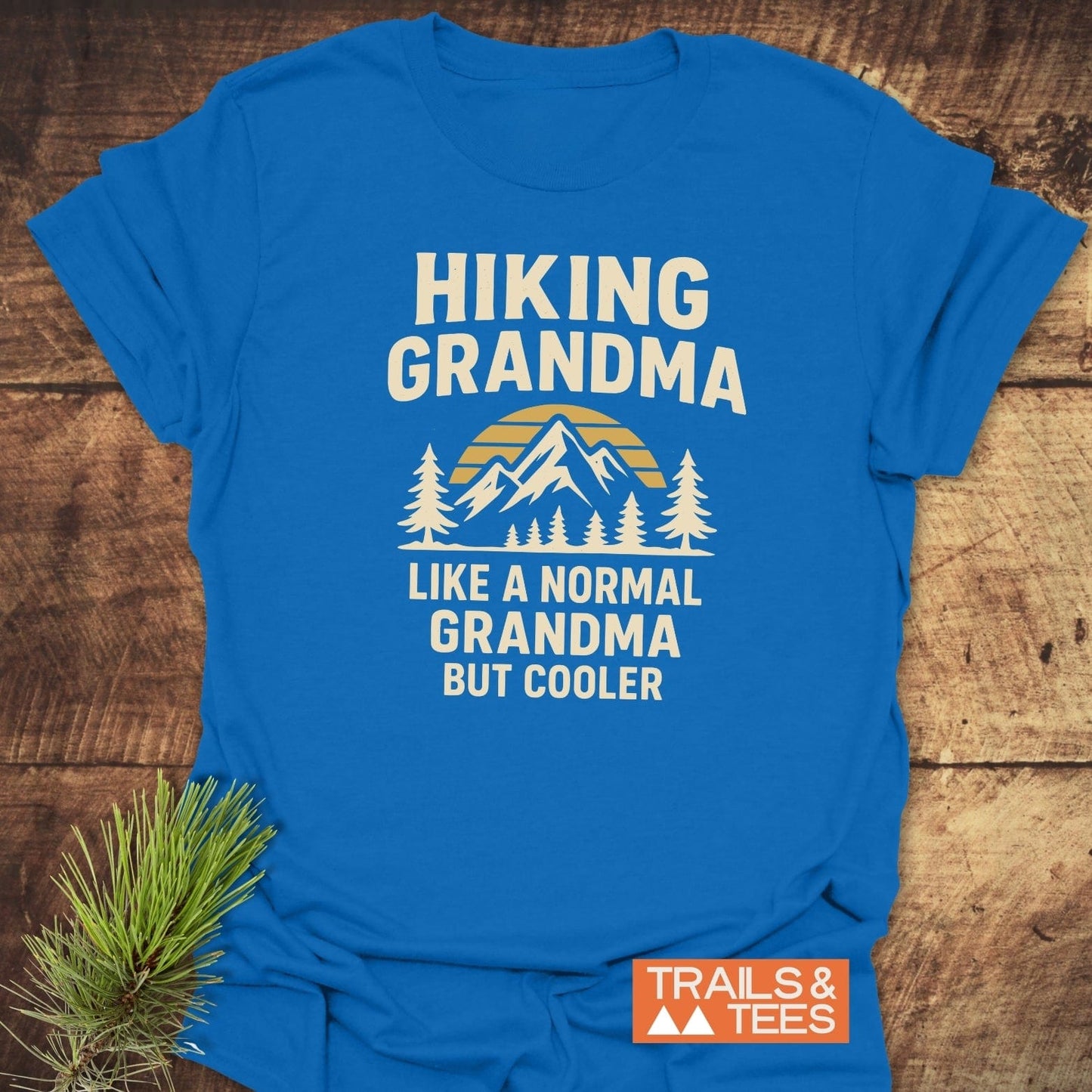 Hiking Grandma Sunrise T-Shirt with a mountain graphic and slogan, laid on wood. Features durable print, soft cotton, pre-shrunk, taped shoulders.