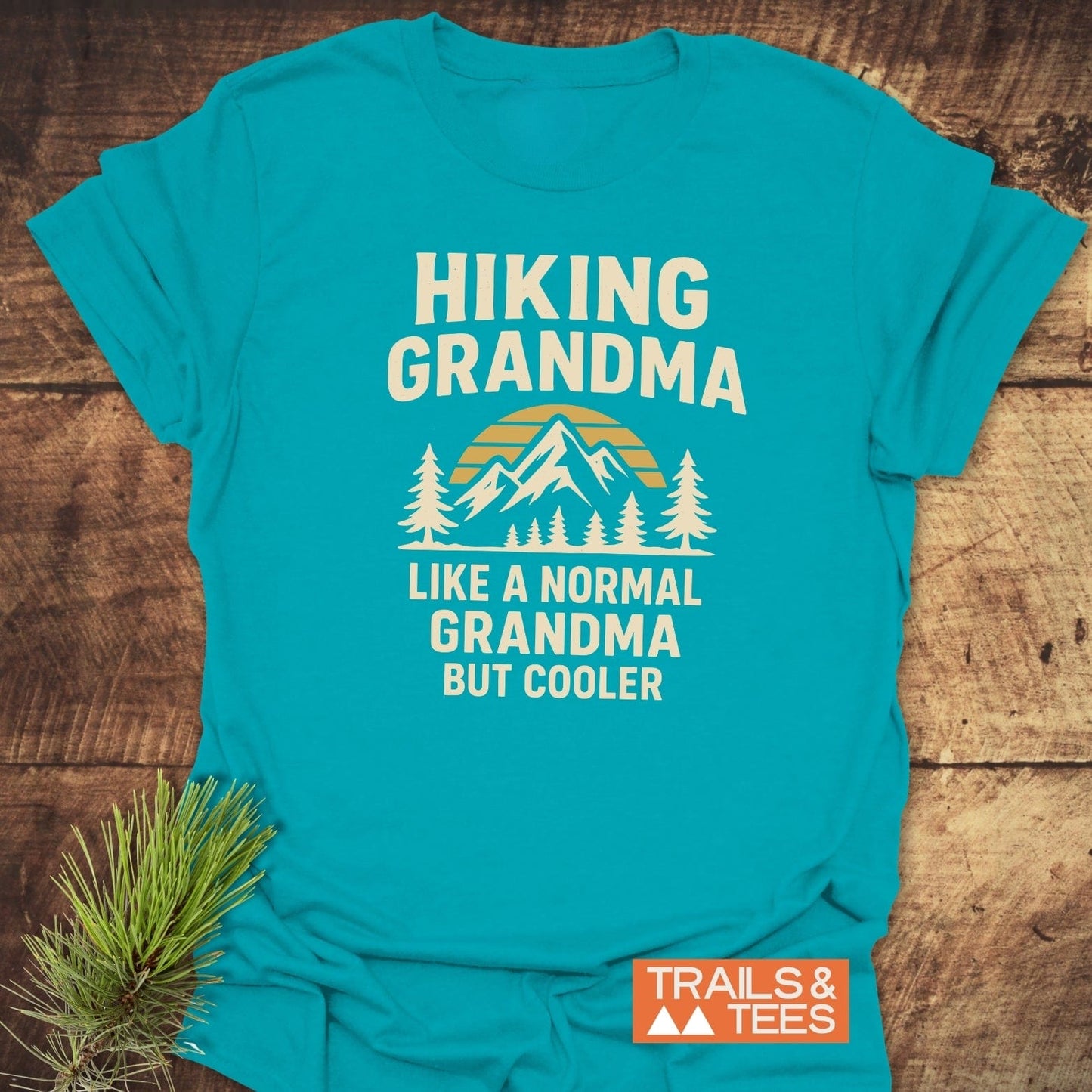 Hiking Grandma Sunrise T-Shirt with a mountain scene and slogan, crafted from soft cotton, laid on wood, featuring a Trails & Tees logo.