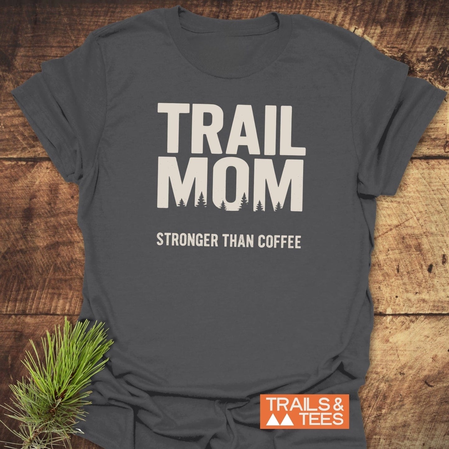 Mom Stronger Than Coffee T-Shirt with tree-line silhouette, cream slogan, and Trails & Tees logo, laid on wood with pine needles for an outdoor-inspired look.