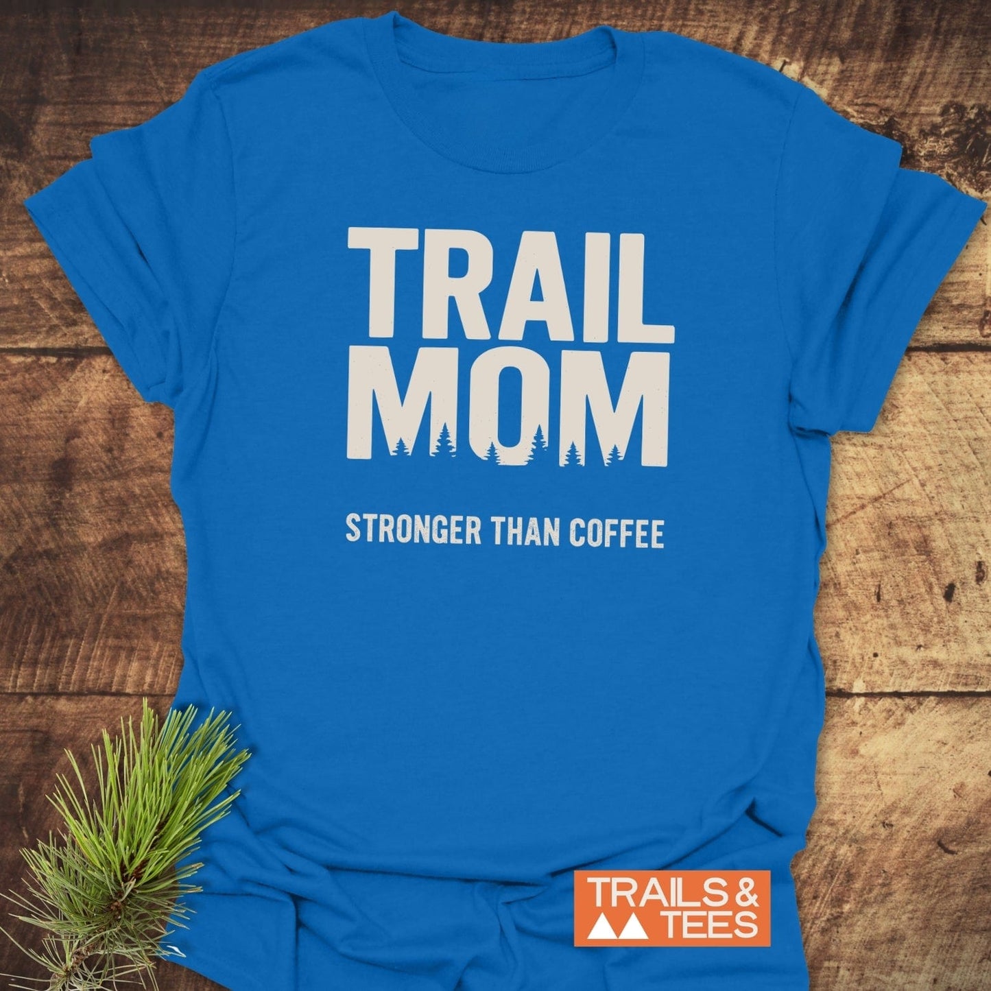 Mom Stronger Than Coffee T-Shirt with 'TRAIL MOM' text and pine-tree details, laid on wood. Soft, pre-shrunk cotton, durable print, designed for outdoor enthusiasts.