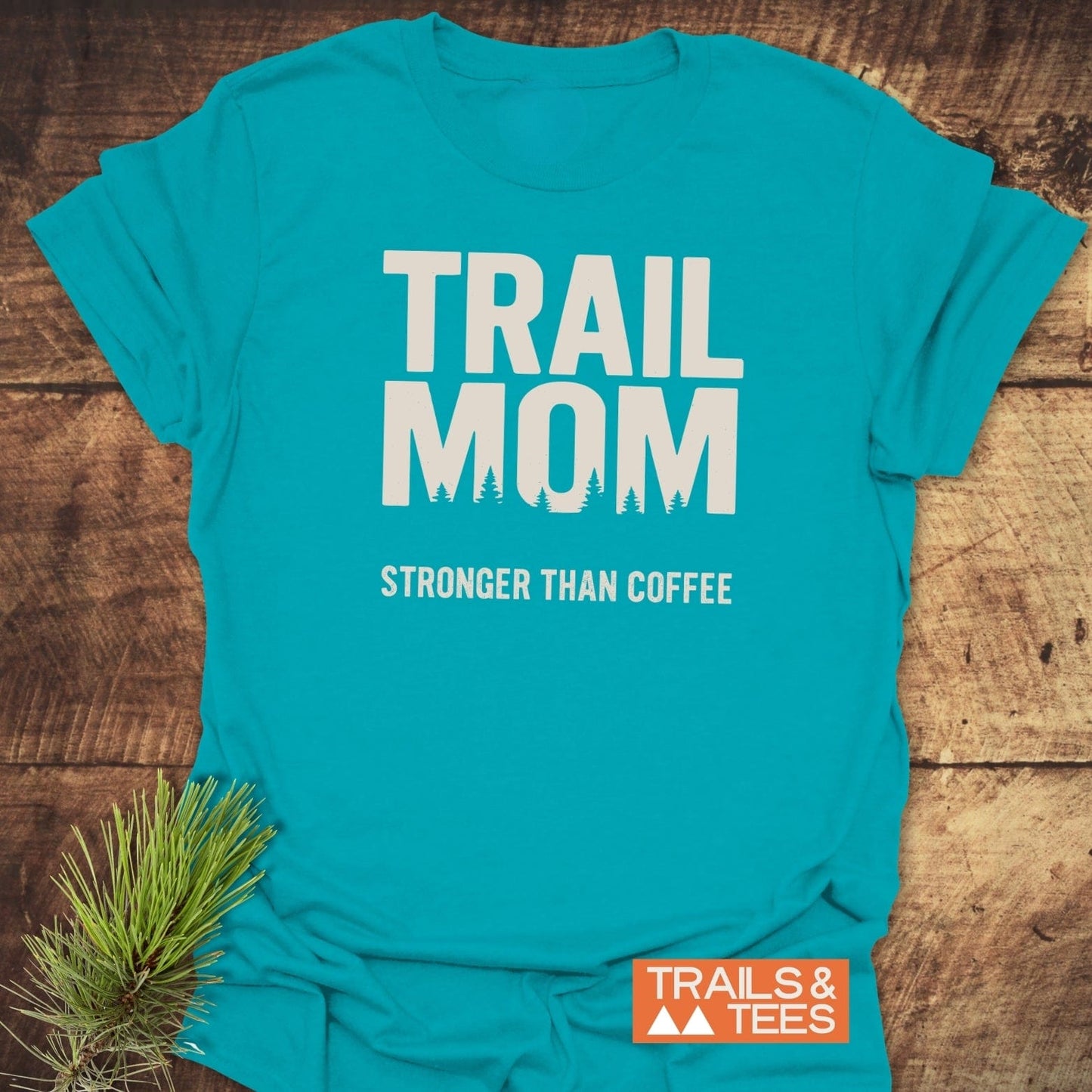 Mom Stronger Than Coffee T-Shirt with bold lettering and pine silhouettes, laid beside a pine sprig, showcases comfort and adventure-ready design.