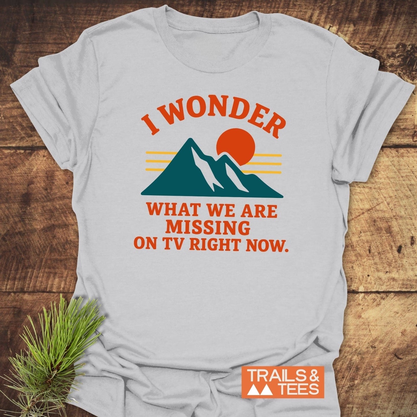 What's On TV Funny T-Shirt featuring teal mountain peaks and red sun graphic with humorous text, laid on wood with pine branch, by Trails & Tees.