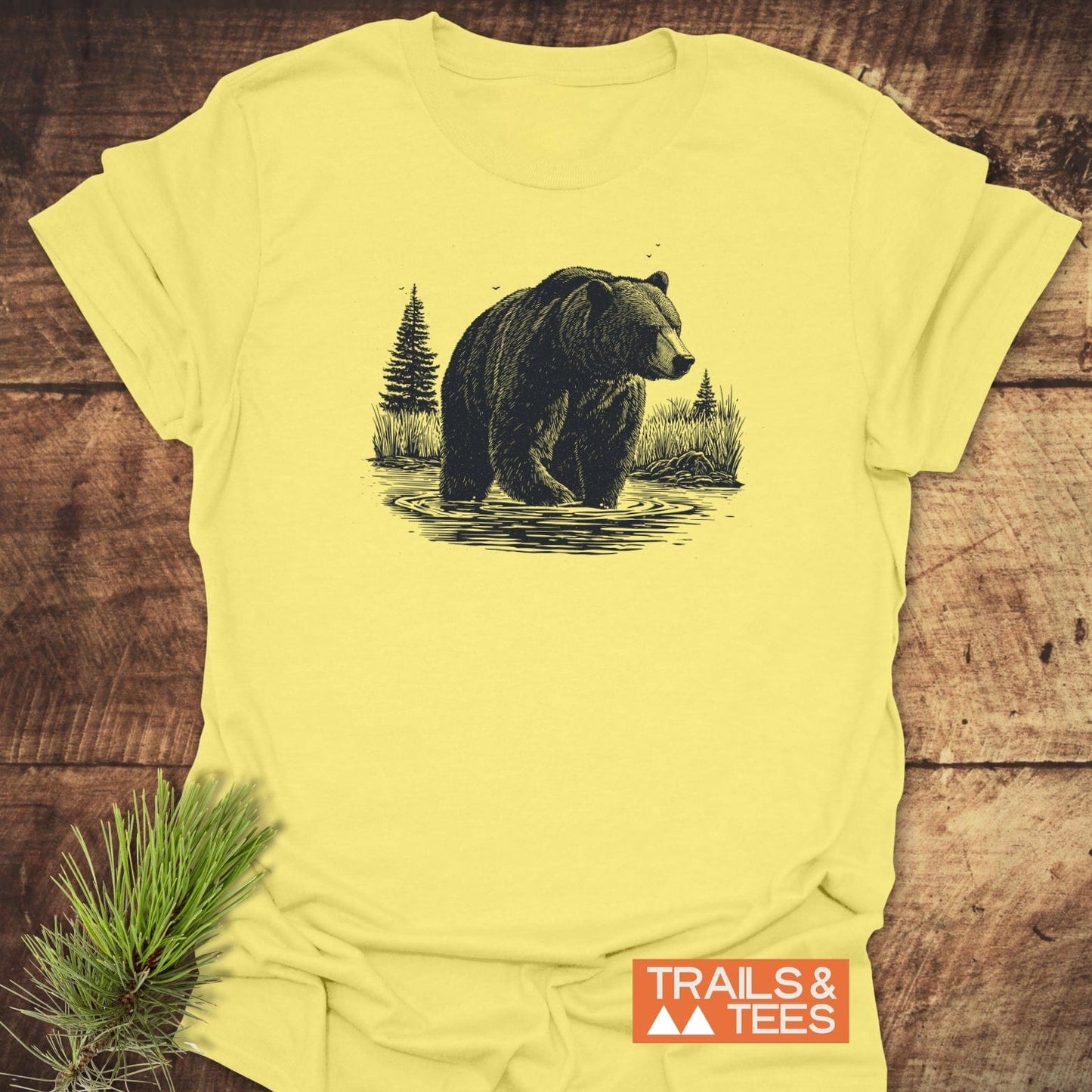 Majestic Grizzly T-Shirt with bear and evergreens graphic on soft, ring-spun cotton, laid on wood, featuring TRAILS & TEES logo.