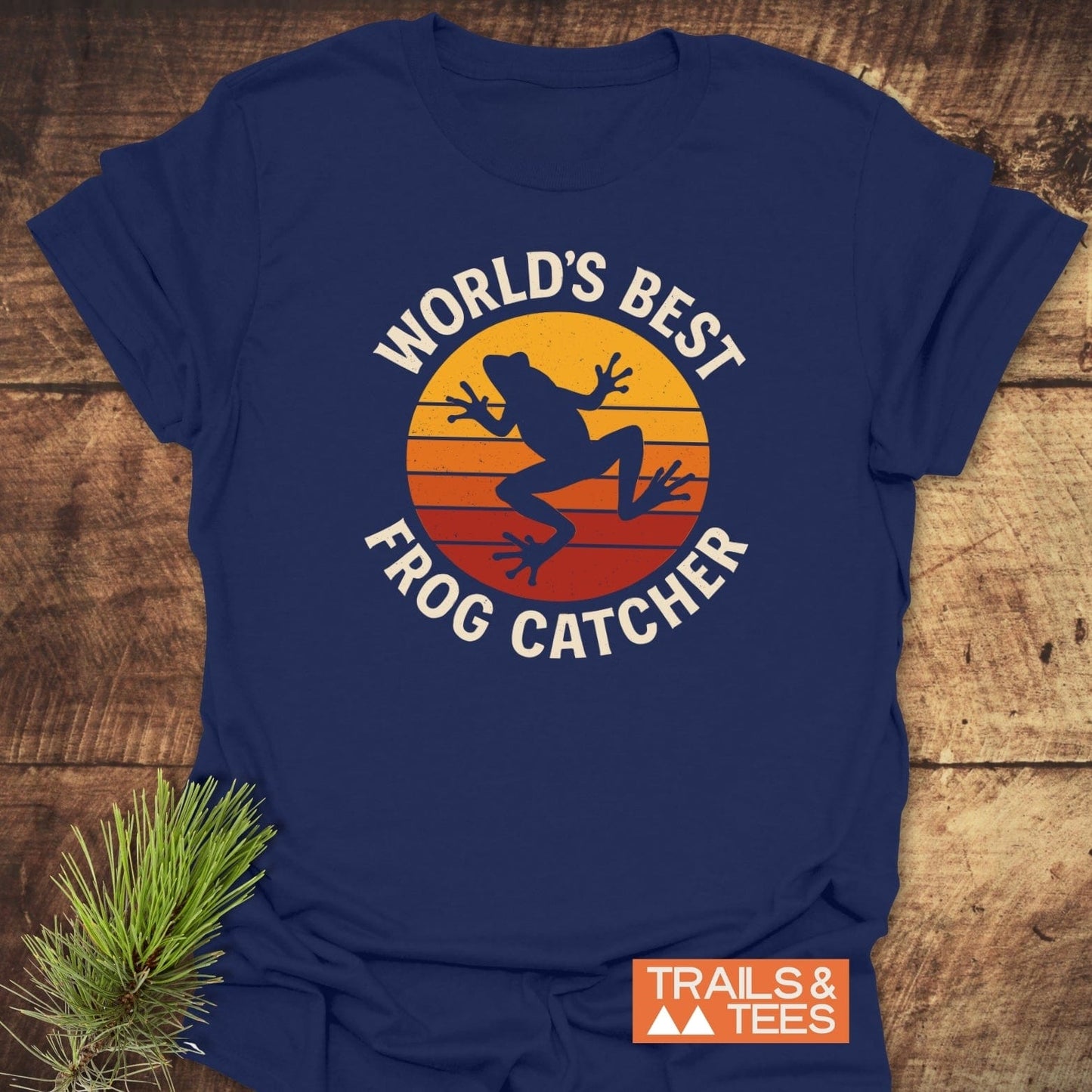Frog Catcher T-Shirt featuring a leaping frog silhouette on a sunset graphic, laid on wood. Made from soft cotton, ideal for outdoor enthusiasts.