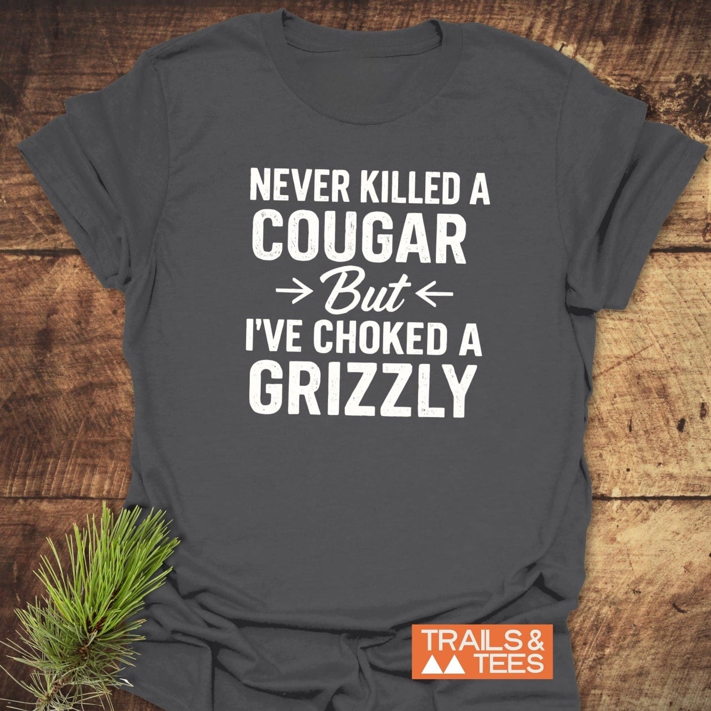 Never Killed A Cougar T-Shirt with bold white text on dark-gray fabric, showcasing outdoor spirit. Laid flat beside pine sprig, Trails & Tees logo visible.