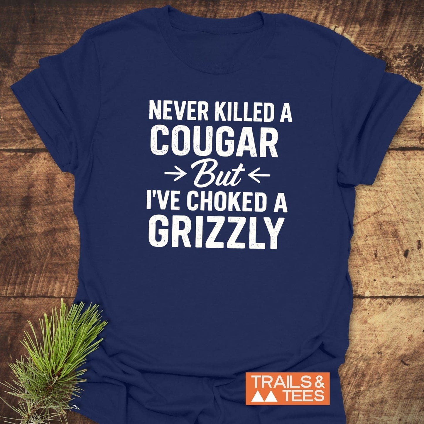 Never Killed A Cougar T-Shirt on wooden surface, featuring bold slogan, Trails & Tees logo, crafted from soft cotton for outdoor adventures.
