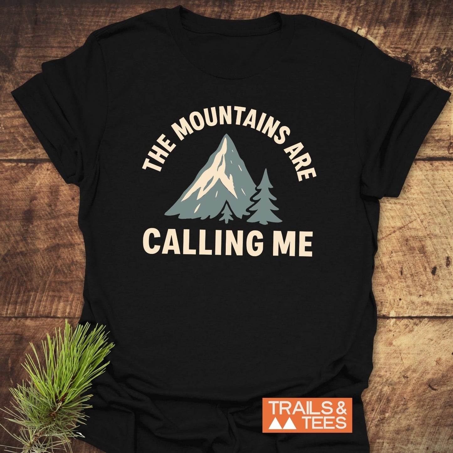 Mountains Calling Me T-Shirt featuring a mountain and trees graphic, with THE MOUNTAINS ARE CALLING ME text, includes an orange TRAILS & TEES logo.