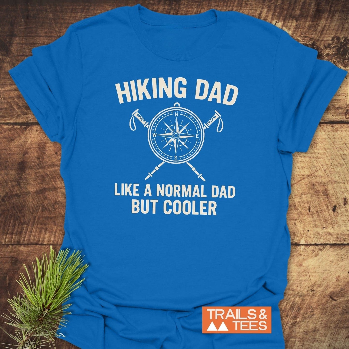 Cool Hiking Dad T-Shirt with compass and trekking poles graphic, laid on wood with pine sprig. Soft, durable cotton for outdoor adventures.