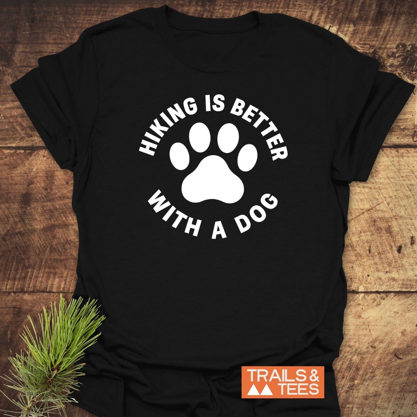 Hiking With Dogs T-Shirt featuring a white dog paw print and slogan on black fabric, symbolizing outdoor adventure. Includes Trails & Tees logo and pine sprig accent.