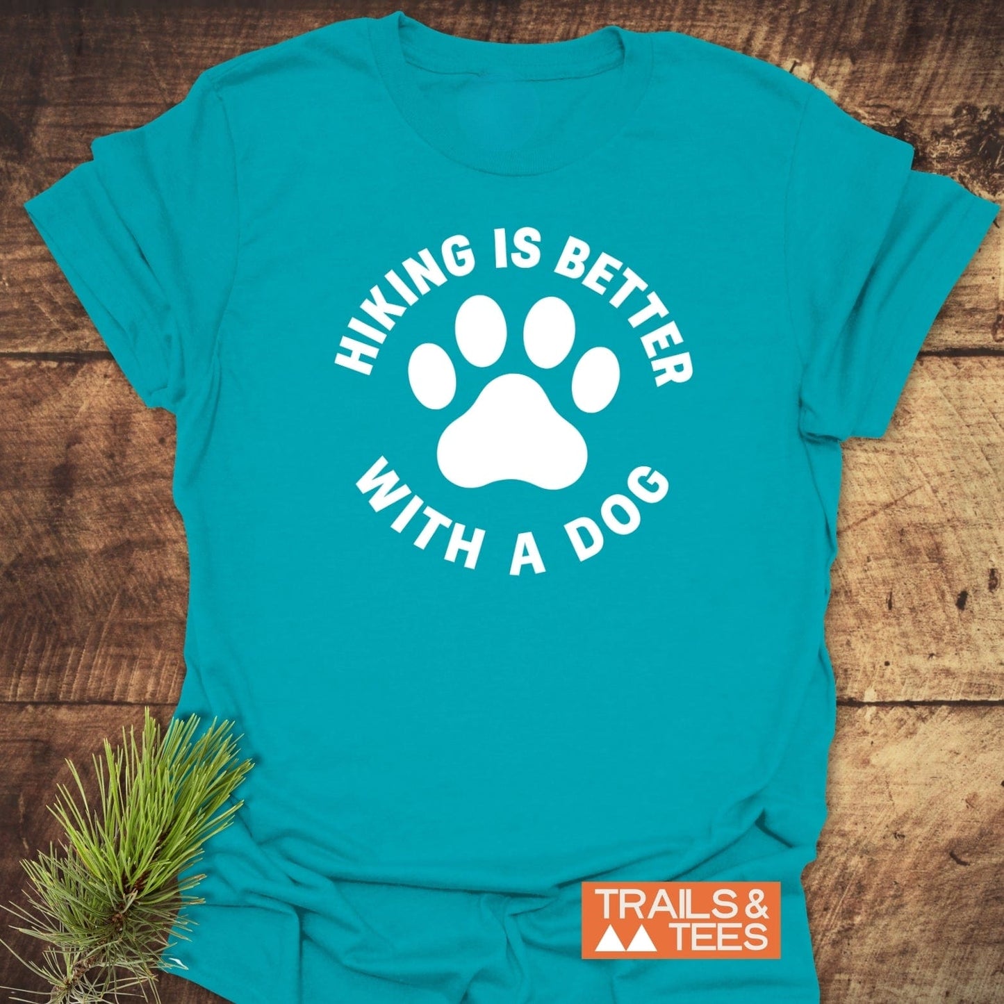 Hiking With Dogs T-Shirt featuring a white dog-paw design and slogan, laid on wood with a rustic pine sprig, highlighting outdoor-inspired comfort and durability.