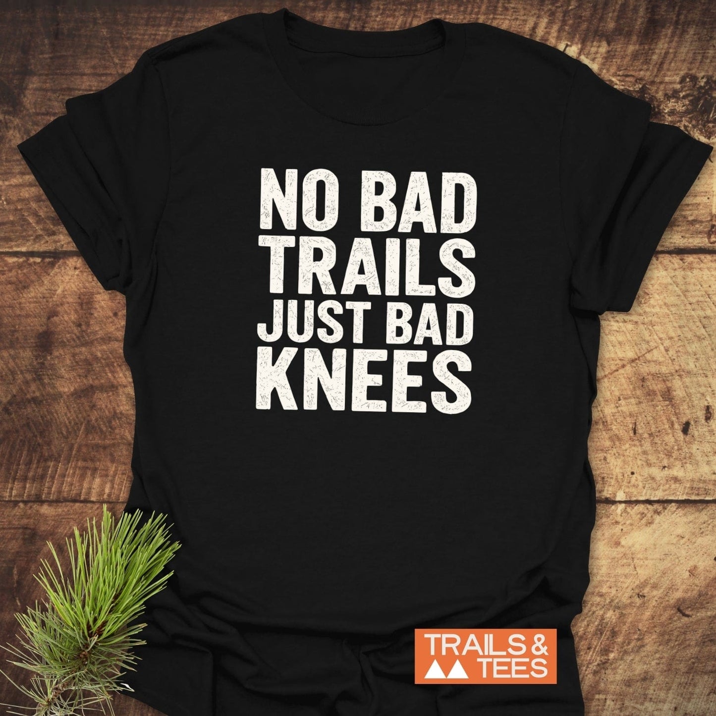 No Bad Trails Funny T-Shirt: black tee with white slogan, No Bad Trails Just Bad Knees, laid flat, featuring TRAILS & TEES logo and pine sprig accent.