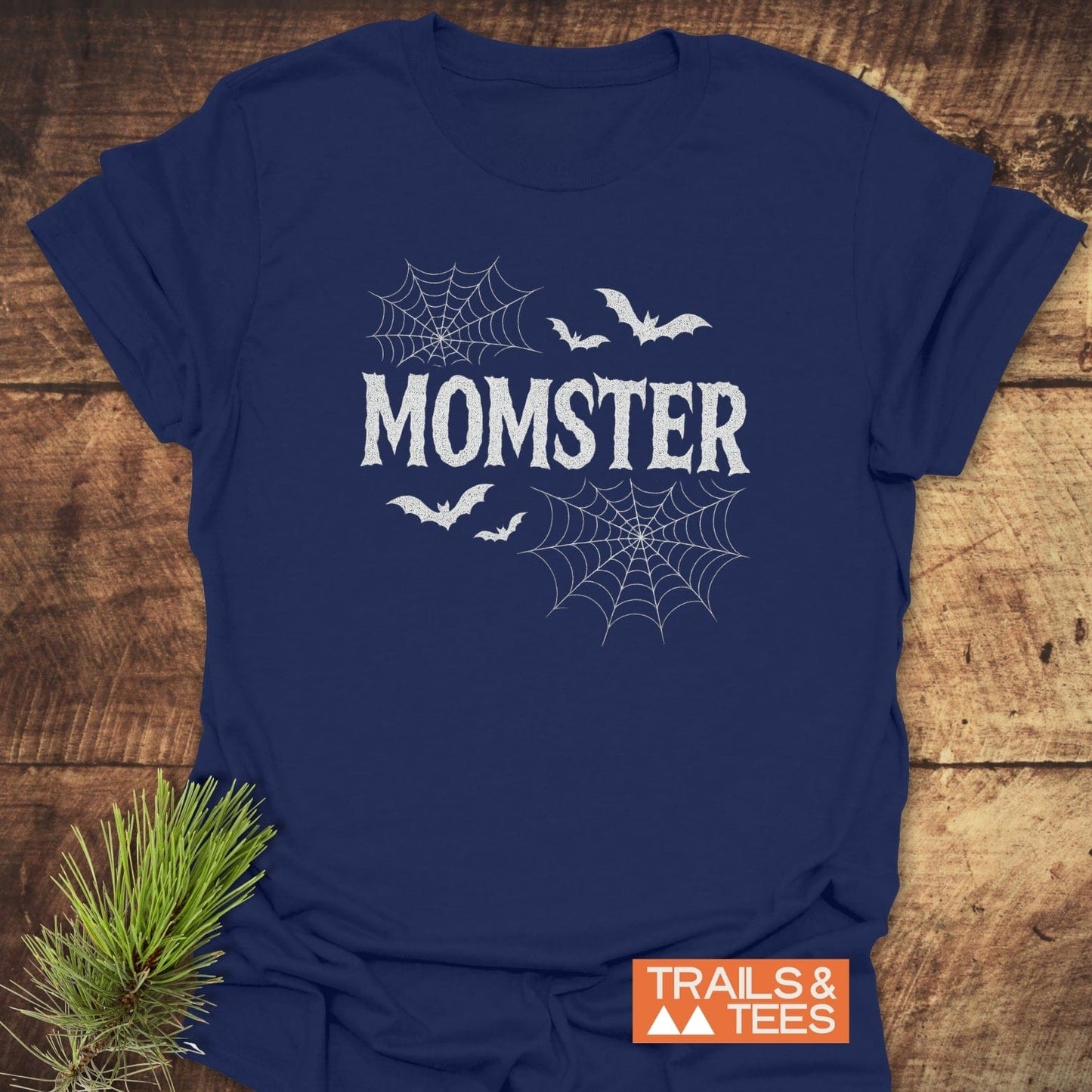 Navy-blue Momster Halloween T-Shirt with spooky font, web graphics, and bats, showcasing Trail-tested comfort and durability. Featuring a Trails & Tees logo and pine sprig accent.