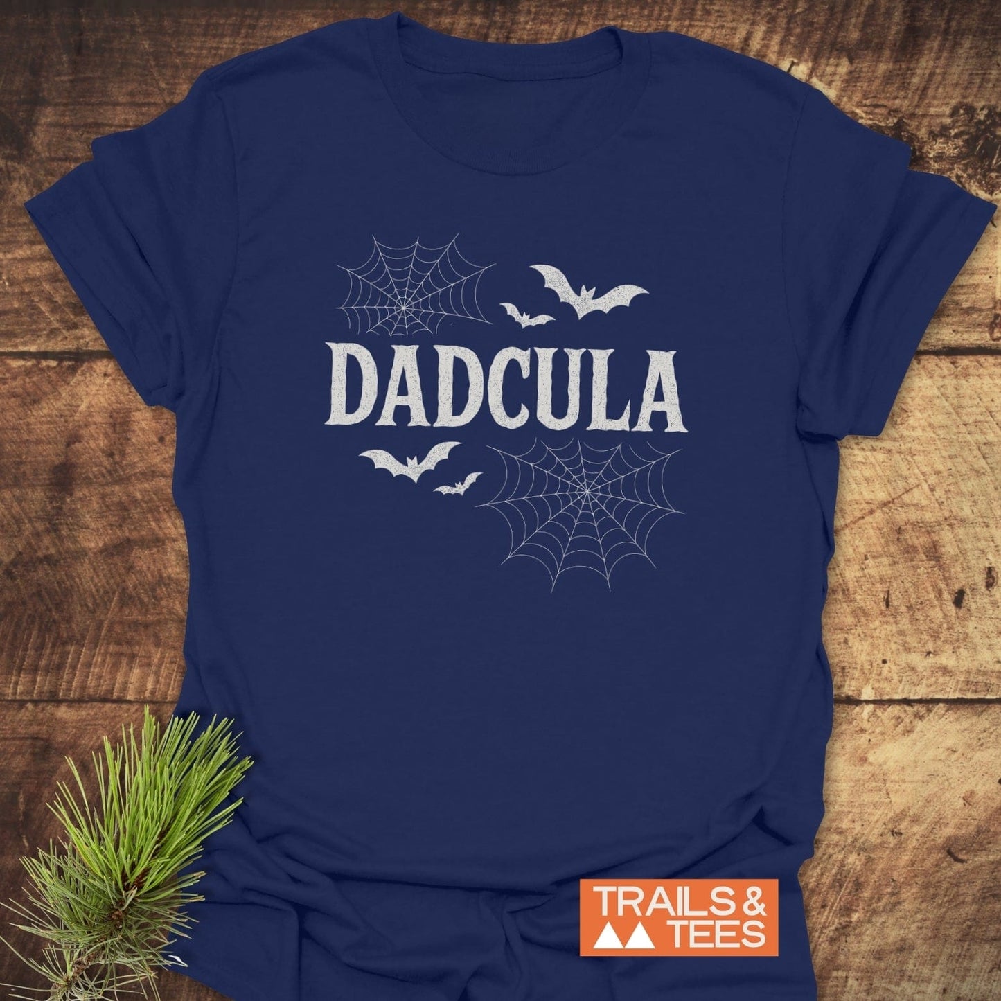 Dadcula Halloween T-Shirt featuring distressed lettering, spider-web graphics, and bat silhouettes, emphasizing comfort and durability for outdoor enthusiasts. Includes Trails & Tees logo and pine sprig accent.