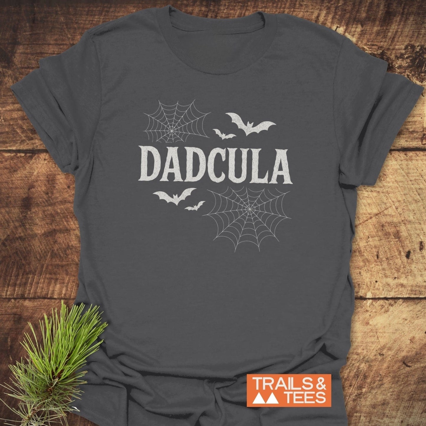 Charcoal-gray DADCULA Halloween T-shirt with bat and spider-web design, laid on wood, featuring durable print and pine needle accent.
