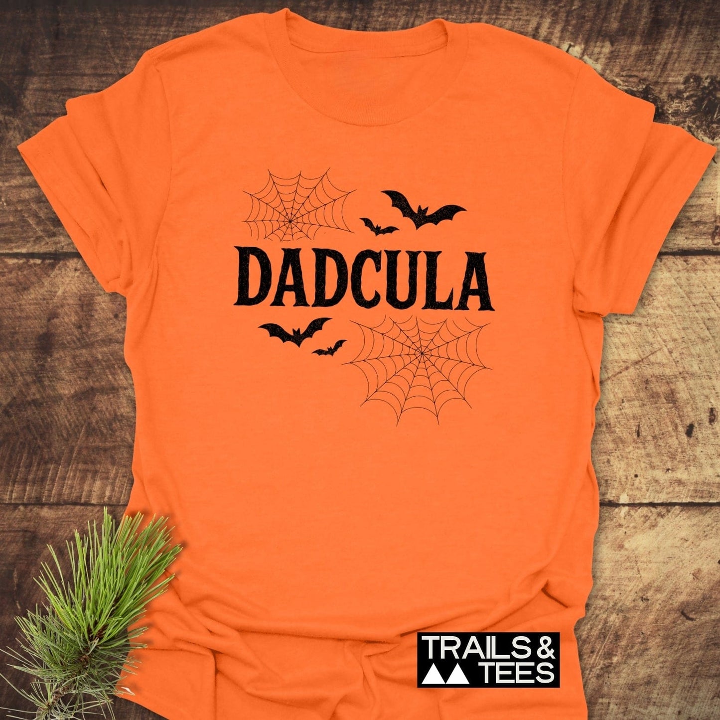 Dadcula Halloween T-Shirt featuring 'DADCULA' text with bats and spider-webs, lying flat with evergreen sprig; soft, durable cotton ideal for outdoor adventures.