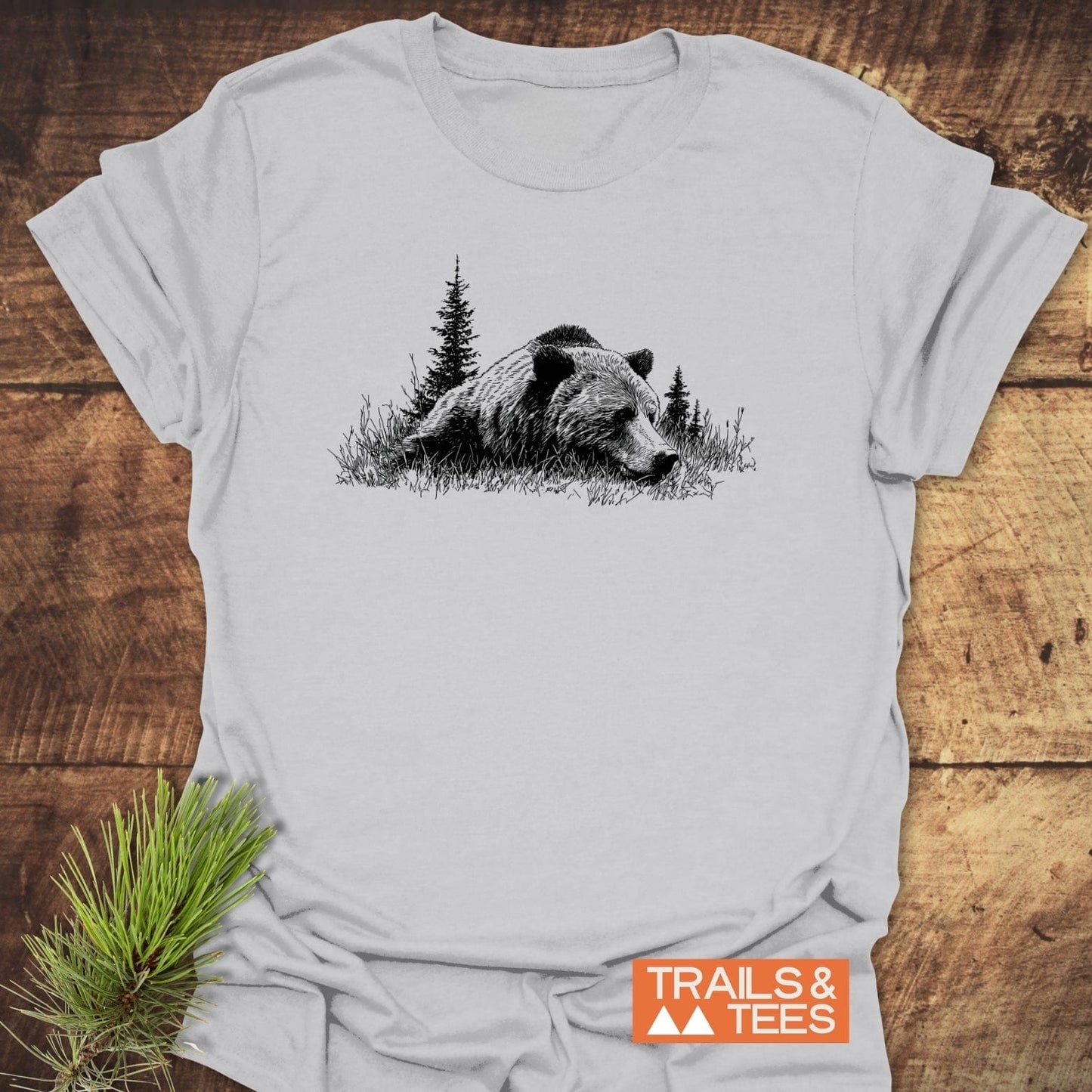 Relaxing Grizzly T-Shirt featuring a black bear and trees illustration on light-grey fabric, with TRAILS & TEES logo, laid on a wooden surface.