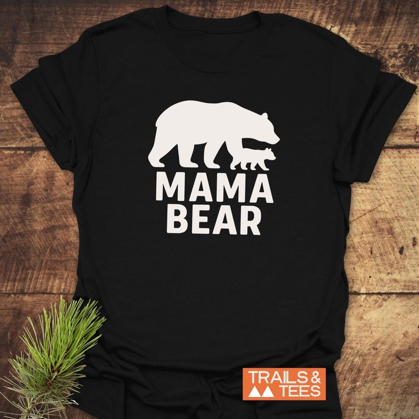 Mama Bear T-Shirt: Black tee with white graphic of mother bear and cub, MAMA BEAR text, styled with pine sprig, Trails & Tees logo visible.