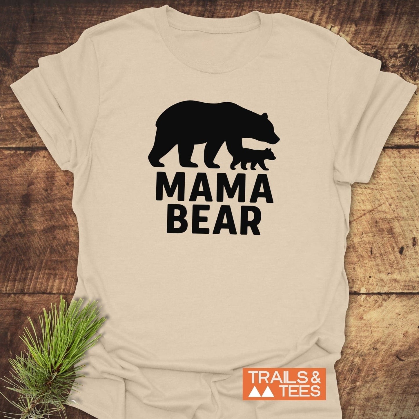 Mama Bear T-Shirt with black bear silhouette, “MAMA BEAR” text, and Trails & Tees logo, laid on rustic wood with pine sprig, showcasing outdoor-inspired design.