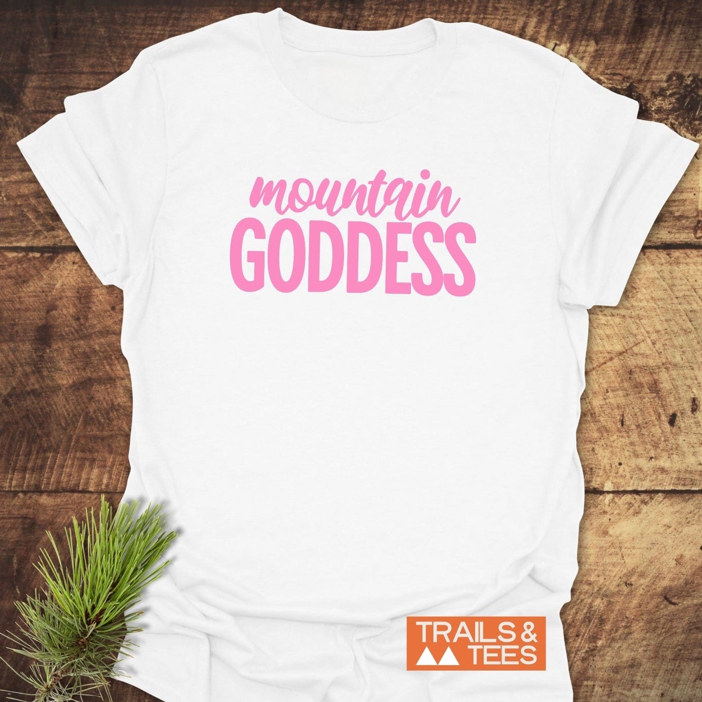 Mountain Goddess T-Shirt featuring pink lettering, displayed on wooden surface, highlights soft ring-spun cotton and durable design with Trails & Tees logo visible.