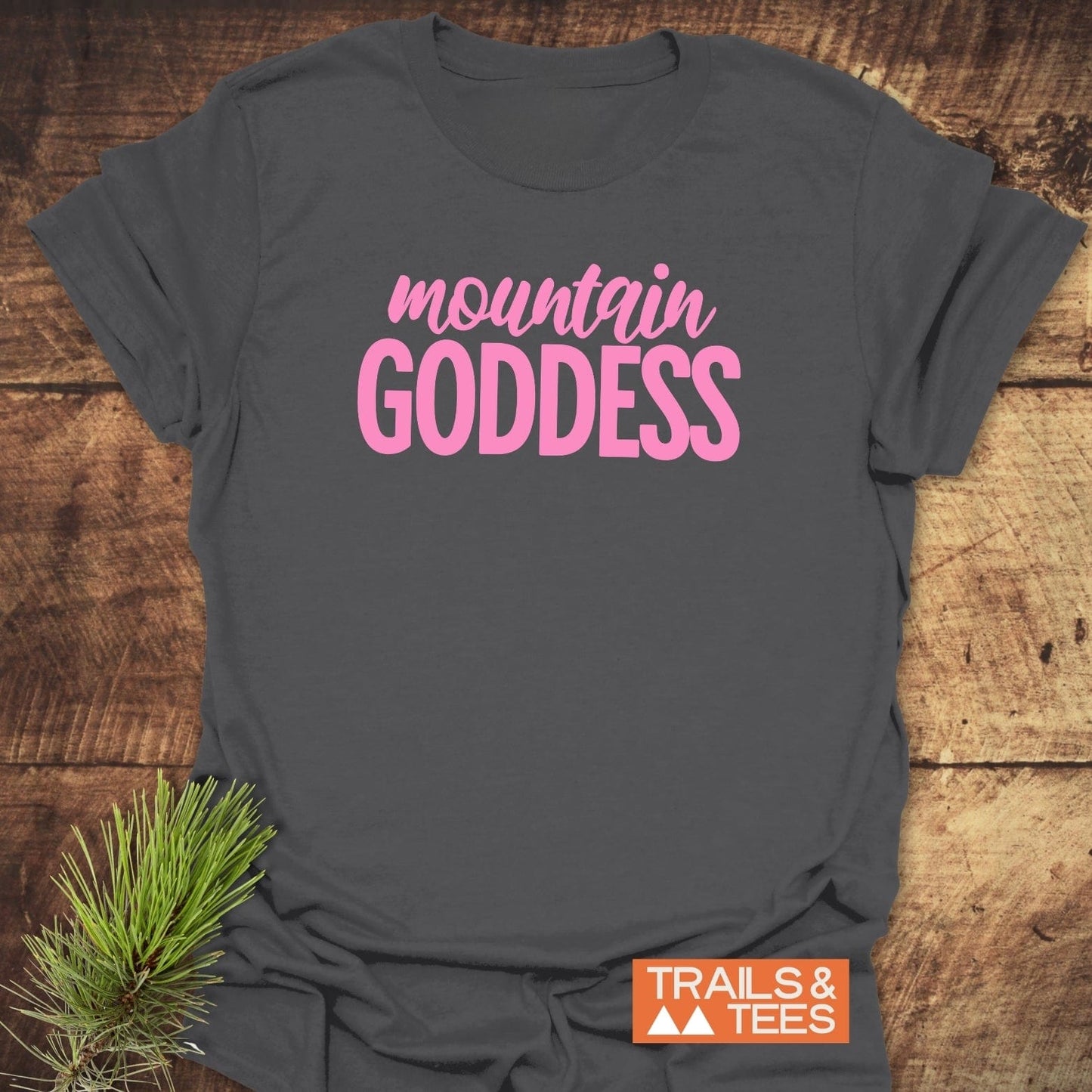 Mountain Goddess T-Shirt featuring bold pink text on soft, dark-gray cotton; ideal for outdoor enthusiasts. Includes Trails & Tees logo and pine sprig accent.