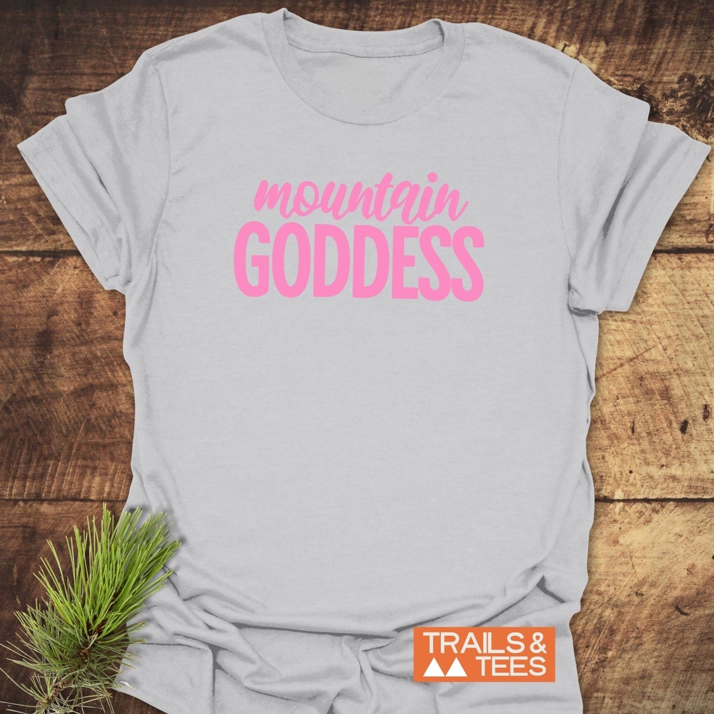 Mountain Goddess T-Shirt featuring bold pink lettering and TRAILS & TEES logo, laid flat with a pine sprig accent, crafted for outdoor enthusiasts.