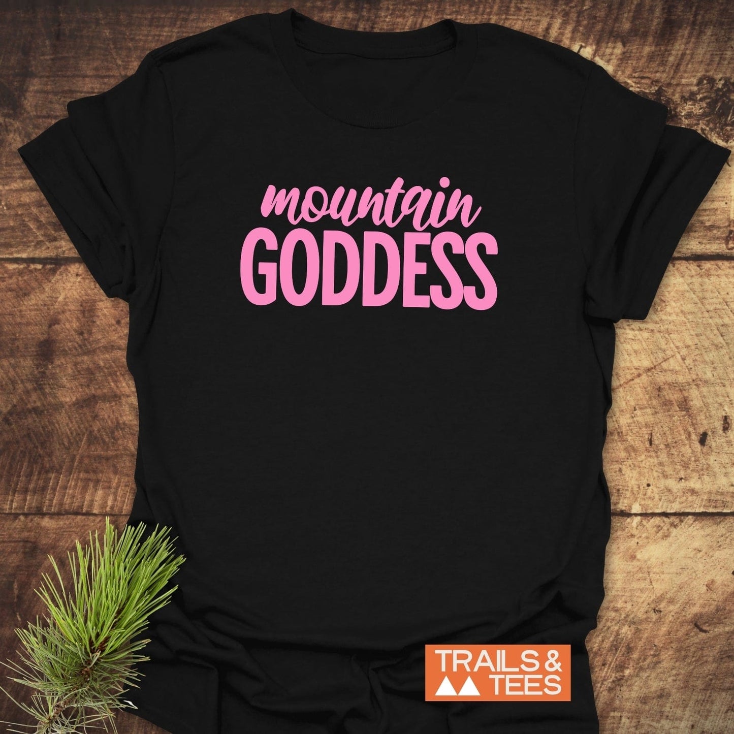 Black Mountain Goddess T-shirt with pink lettering, featuring a small Trails & Tees logo, laid flat on a wooden surface with pine sprig.