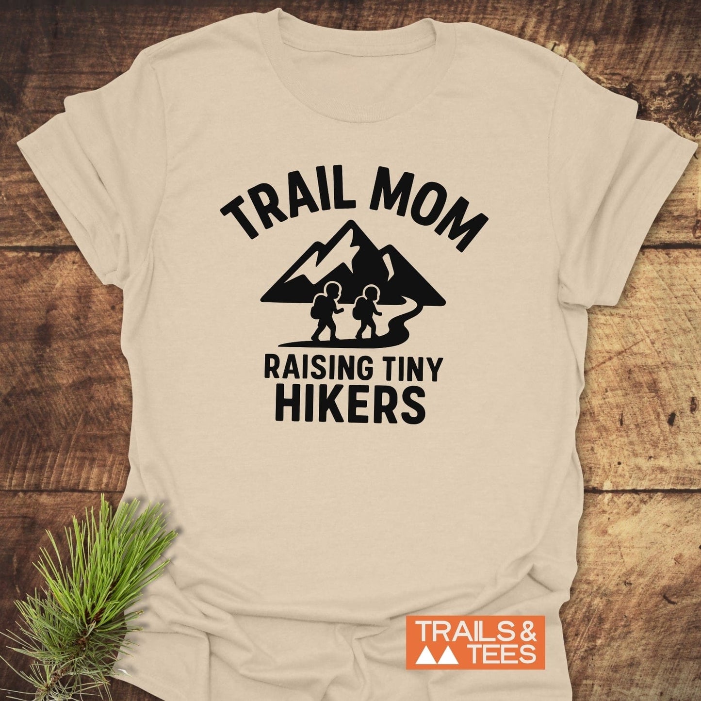Raising Tiny Hikers Mom T-Shirt with TRAIL MOM graphic, mountain silhouette, and slogan. Soft ring-spun cotton, durable print, emphasizes outdoor adventures.