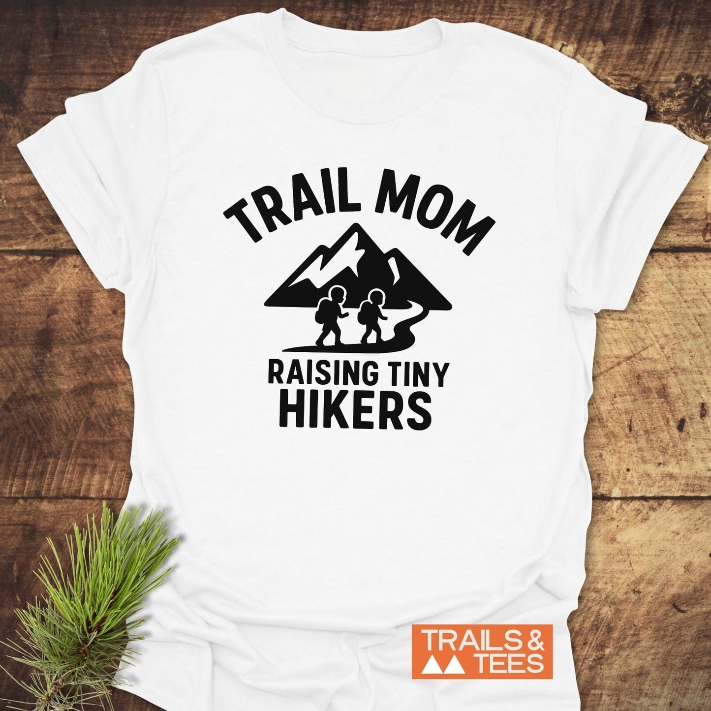 Raising Tiny Hikers Mom T-Shirt featuring black trail graphic and text, laid on wood with pine sprig. Comfortable, ring-spun cotton design, ideal for outdoor enthusiasts.