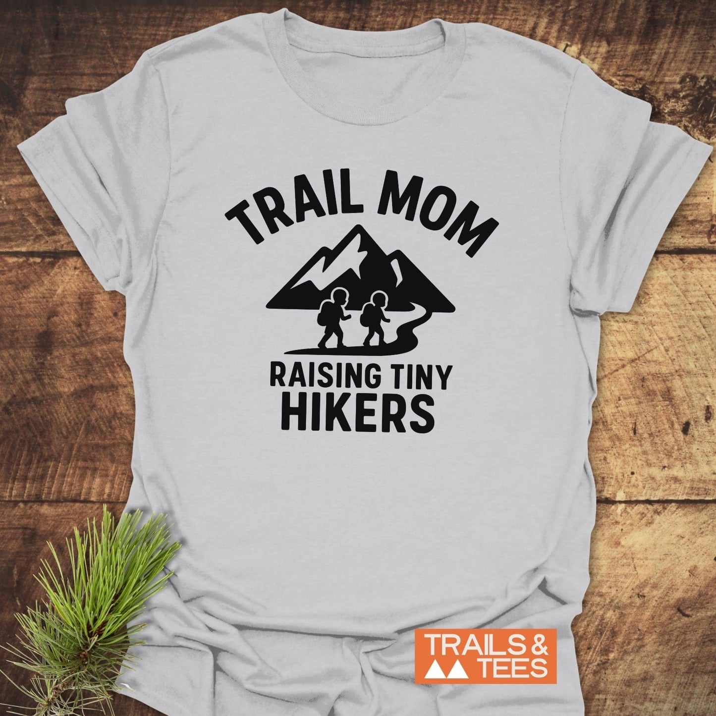 Raising Tiny Hikers Mom T-Shirt with bold graphic of hikers and mountains, laid flat with a pine sprig. Made from soft, durable ring-spun cotton.