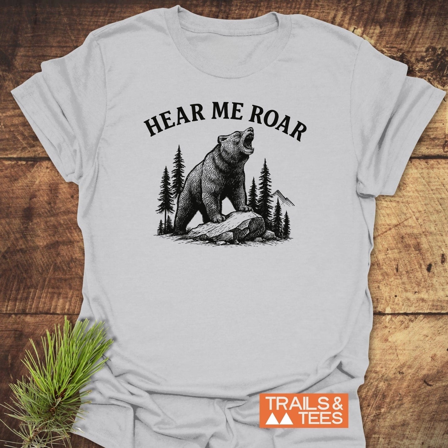 Hear Me Roar T-Shirt featuring a black graphic of a roaring bear on a rock with trees and mountains, laid on wood, beside a pine sprig.