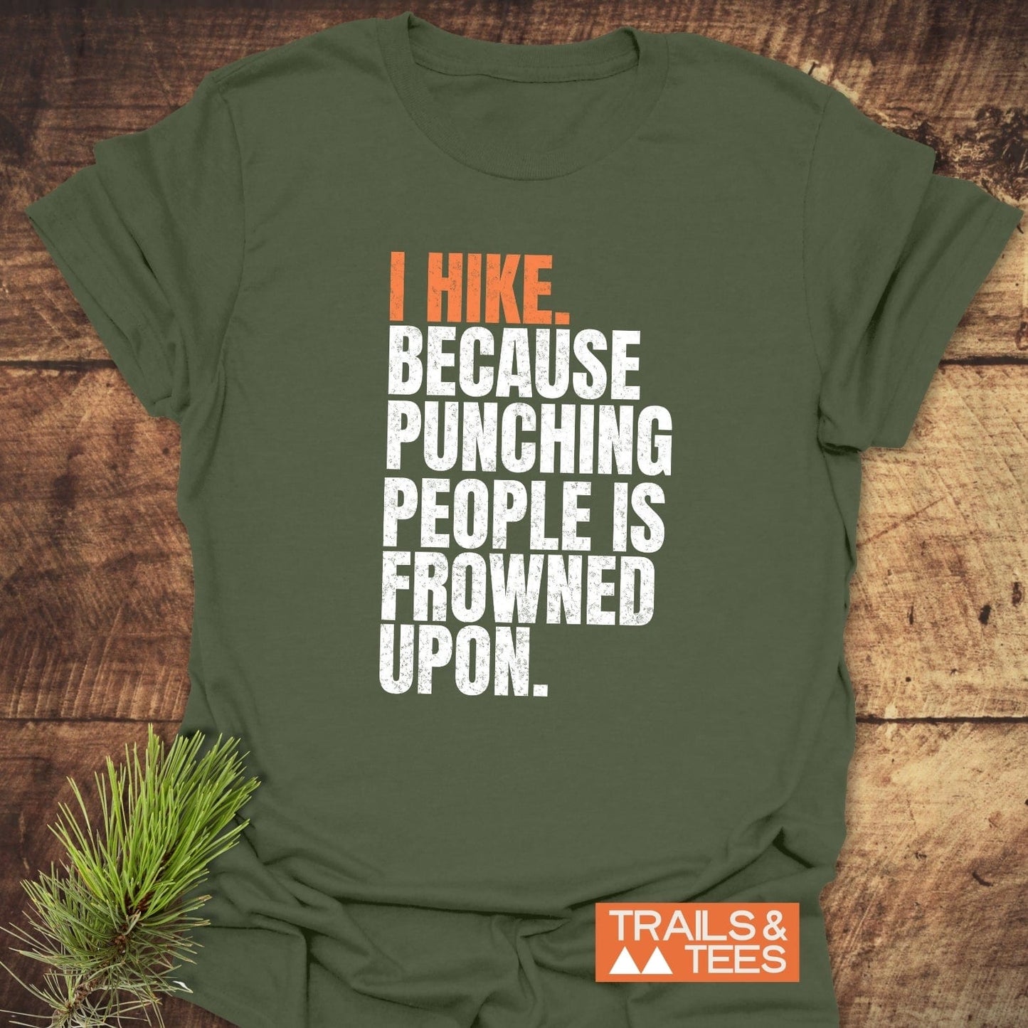 Olive-green I Hike. Funny T-Shirt with bold orange-white slogan, featuring TRAILS & TEES logo. Displayed on wood with pine sprig, highlighting outdoor adventure spirit.