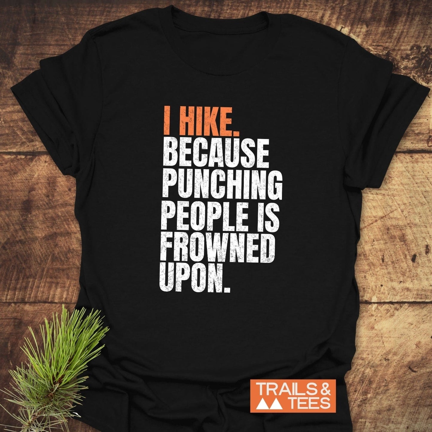 Black I Hike. Funny T-Shirt on wood, featuring a humorous slogan in white and orange. Includes Trails & Tees logo, emphasizing outdoor adventure and comfort.