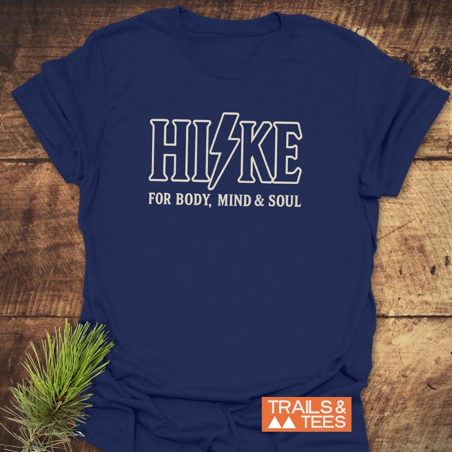 Navy-blue HIKE Tour T-Shirt on wood, featuring a lightning bolt I and slogan, crafted from soft cotton with durable prints and Trails & Tees logo.