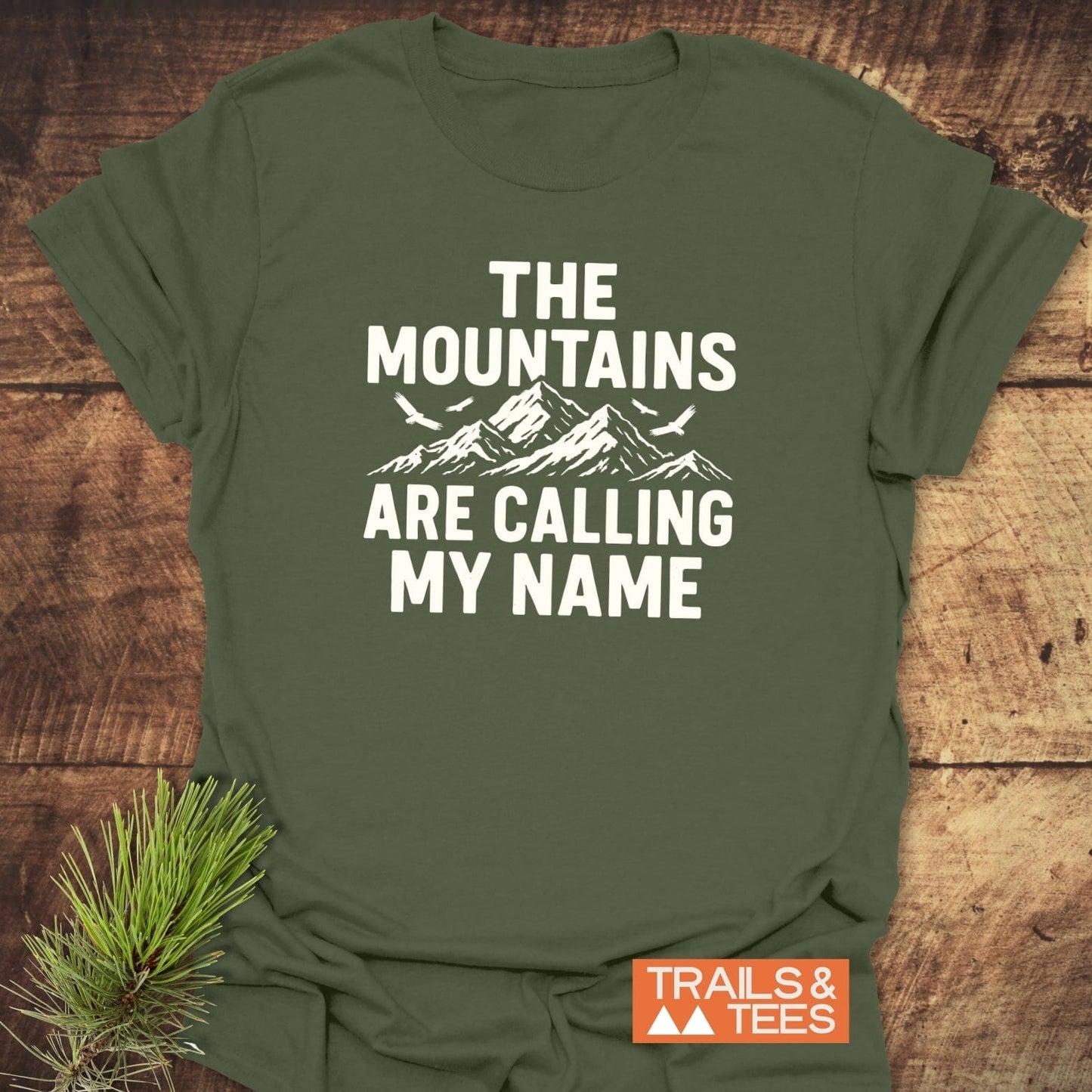 Calling My Name T-Shirt featuring mountain and bird graphic, slogan, and Trails & Tees label on soft, army-green cotton, laid on wooden surface with pine sprig.