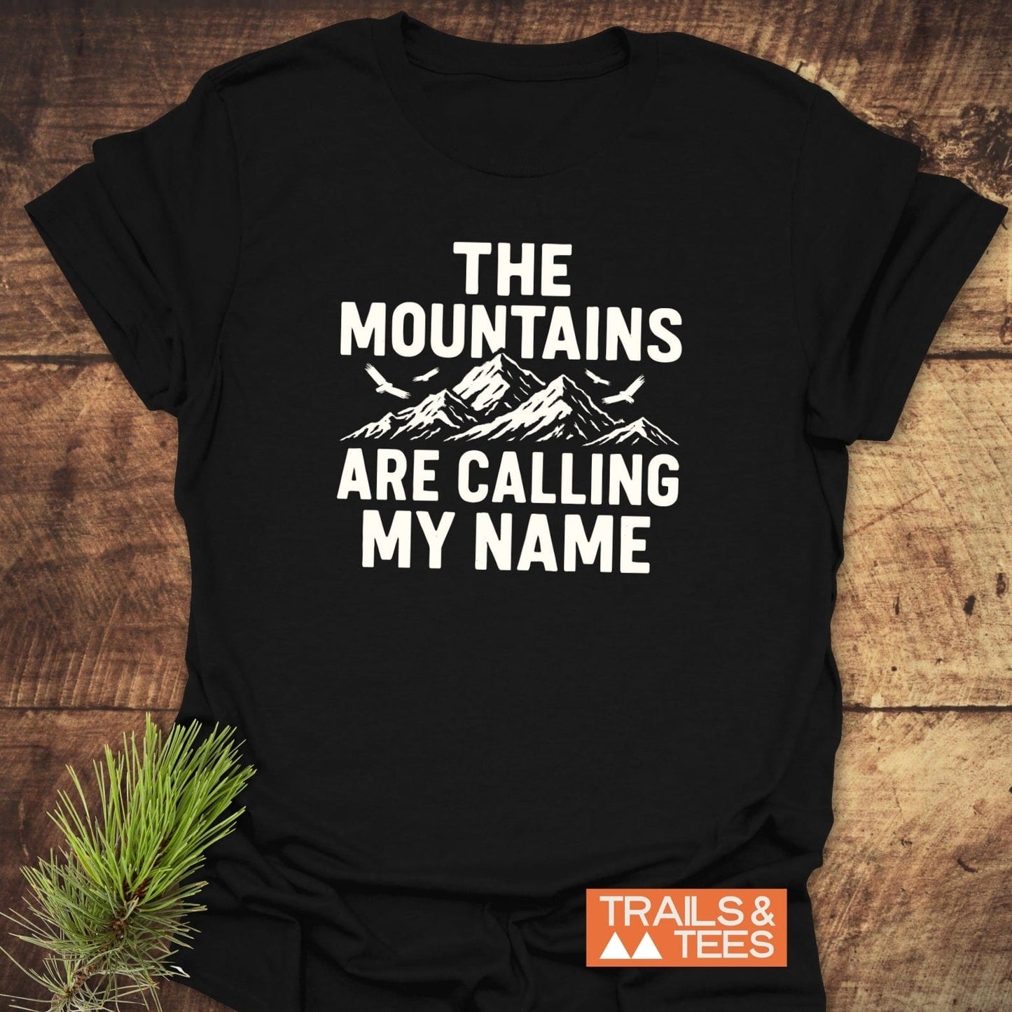 Black 'Calling My Name' T-shirt with mountain graphic, birds, and bold text, laid on wood. Features soft, durable cotton for outdoor enthusiasts.